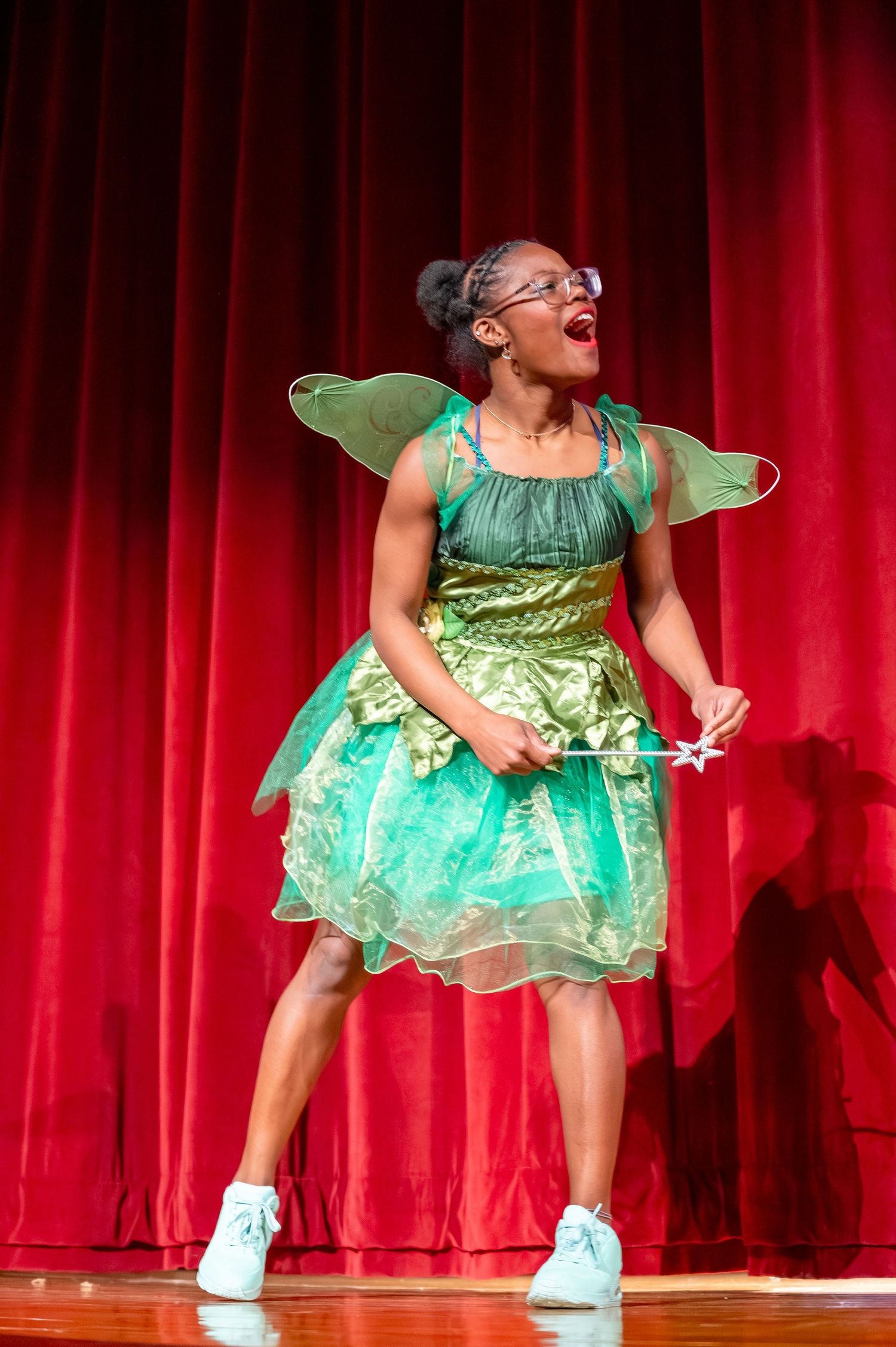 Performer in sparkly green fairy costume and white sneakers dances energetically on stage in front of red curtain.