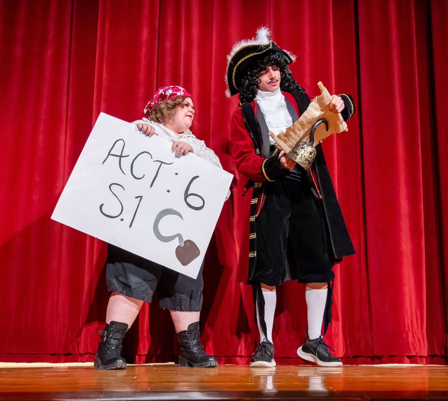 Performers on stage hold up score cards against a red curtain backdrop during a theatrical performance.