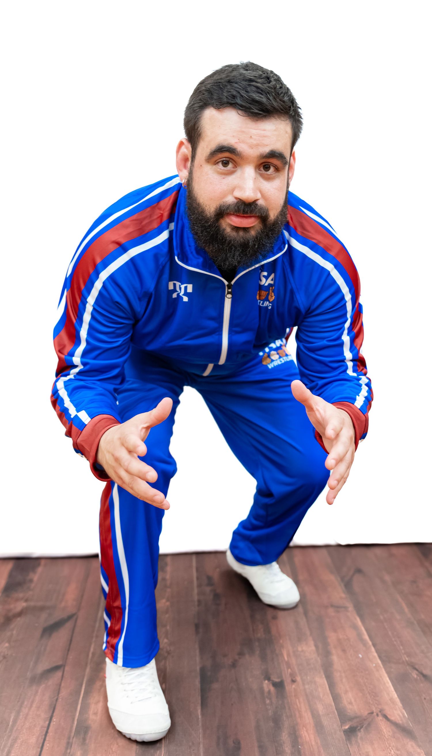 Athlete in blue and red USA Wrestling tracksuit demonstrates dynamic crouching poses on wooden floor.