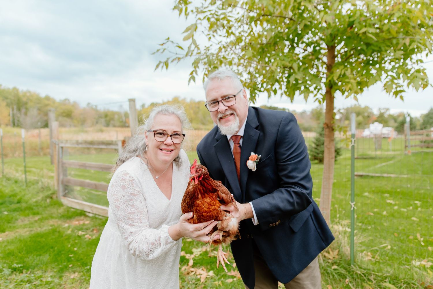 Playful wedding portrait with couple holding a chicken on farm grounds.