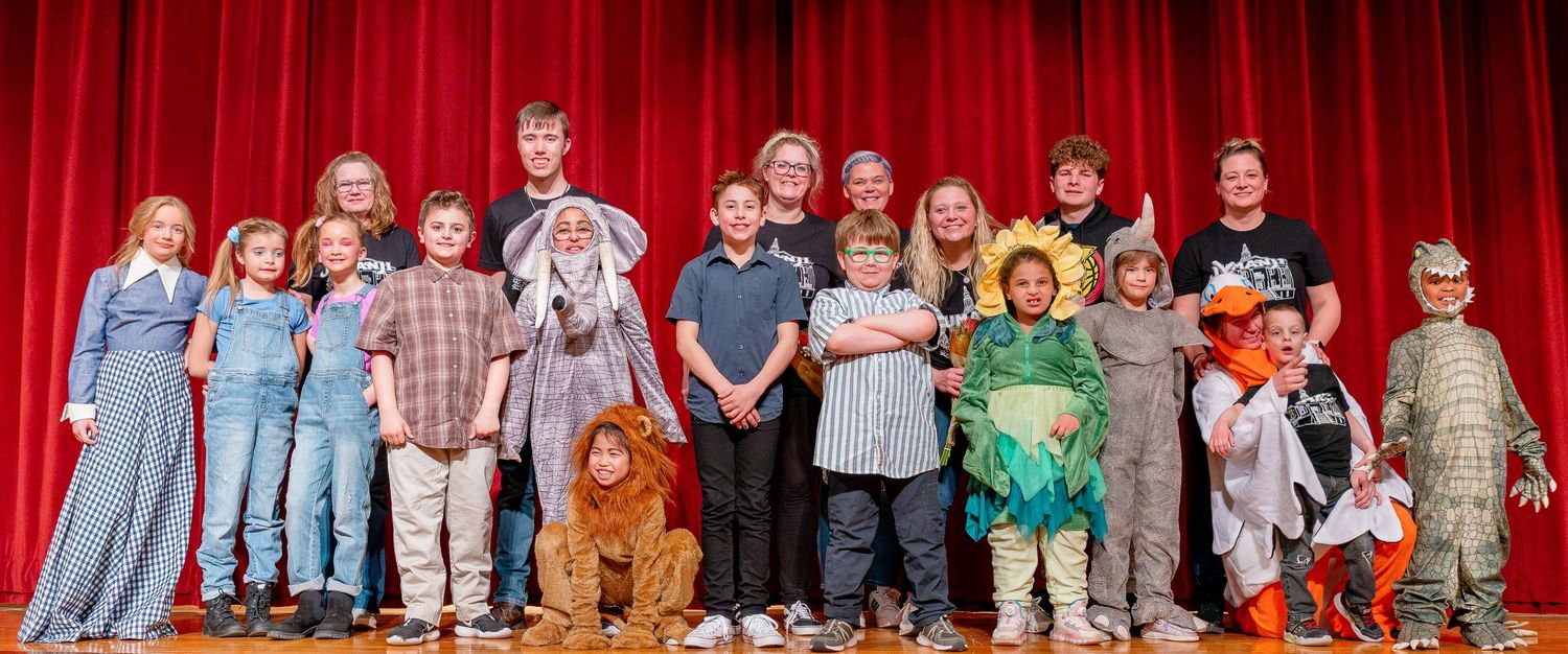 Large group of elementary school students pose on stage with colorful costumes for a theatrical performance.
