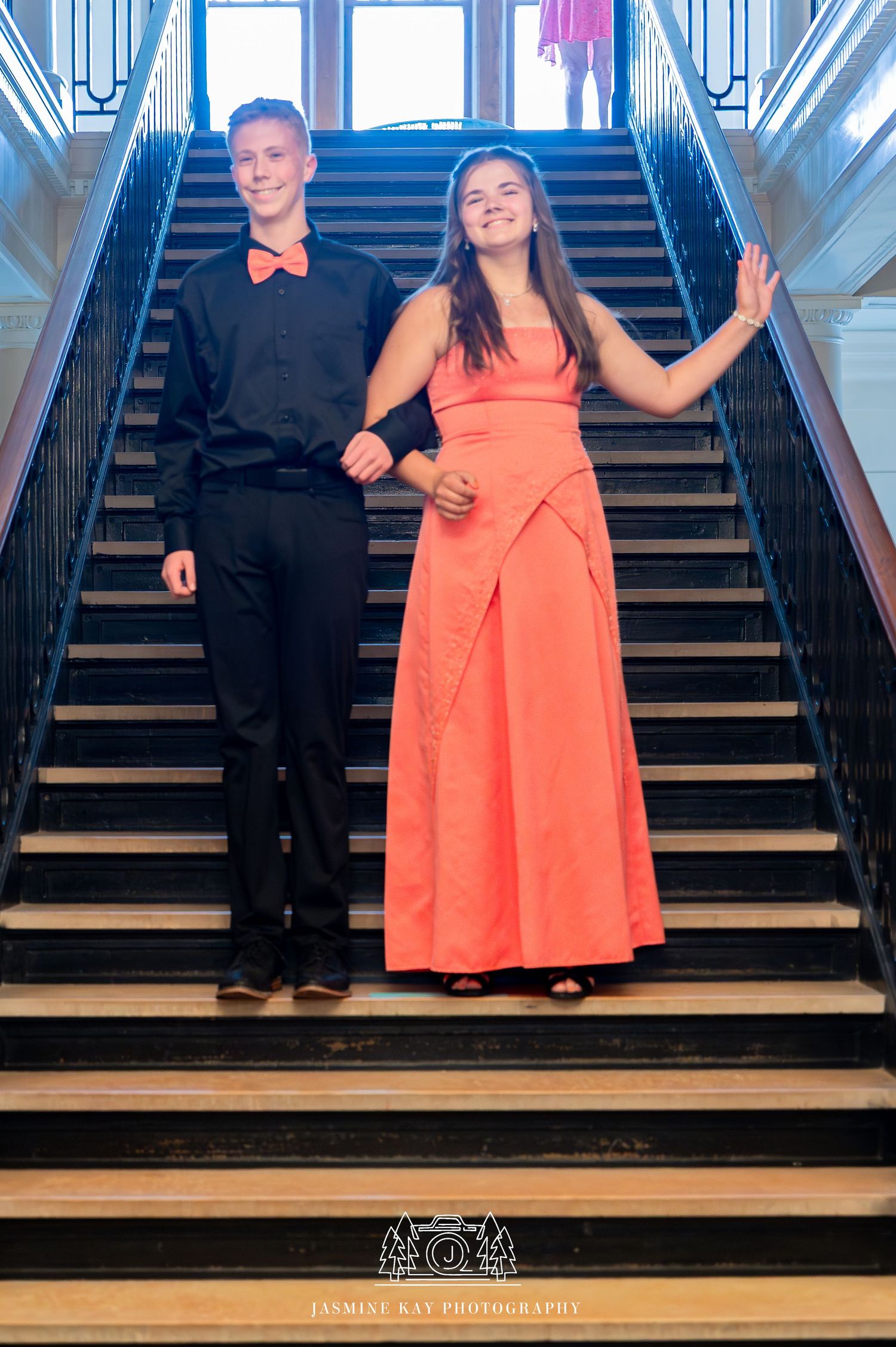 A couple poses on elegant stairs, with one wearing a black suit and bow tie and the other in a coral formal gown.