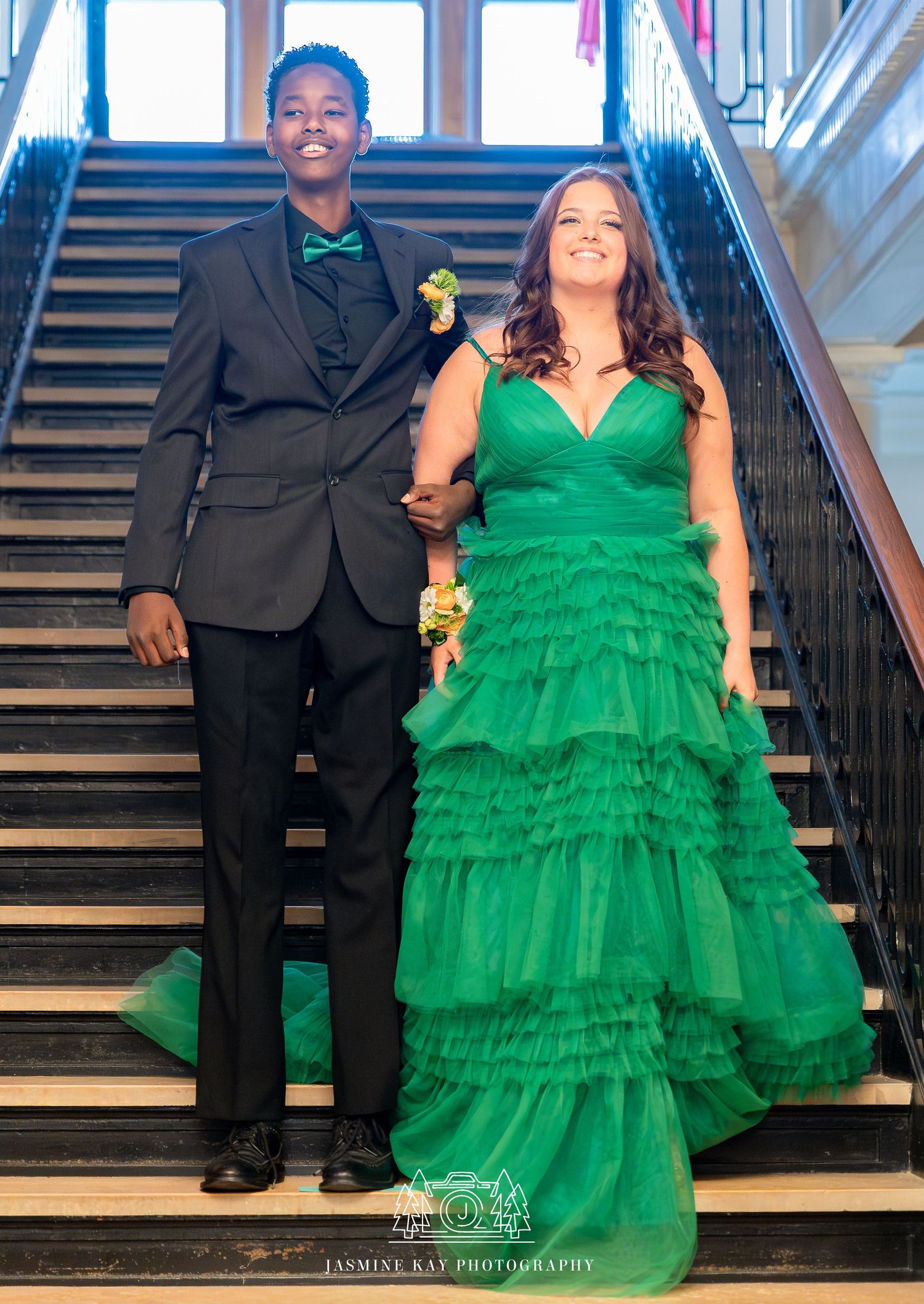 A couple poses on elegant stairs, with the woman wearing a vibrant emerald green tiered tulle gown.