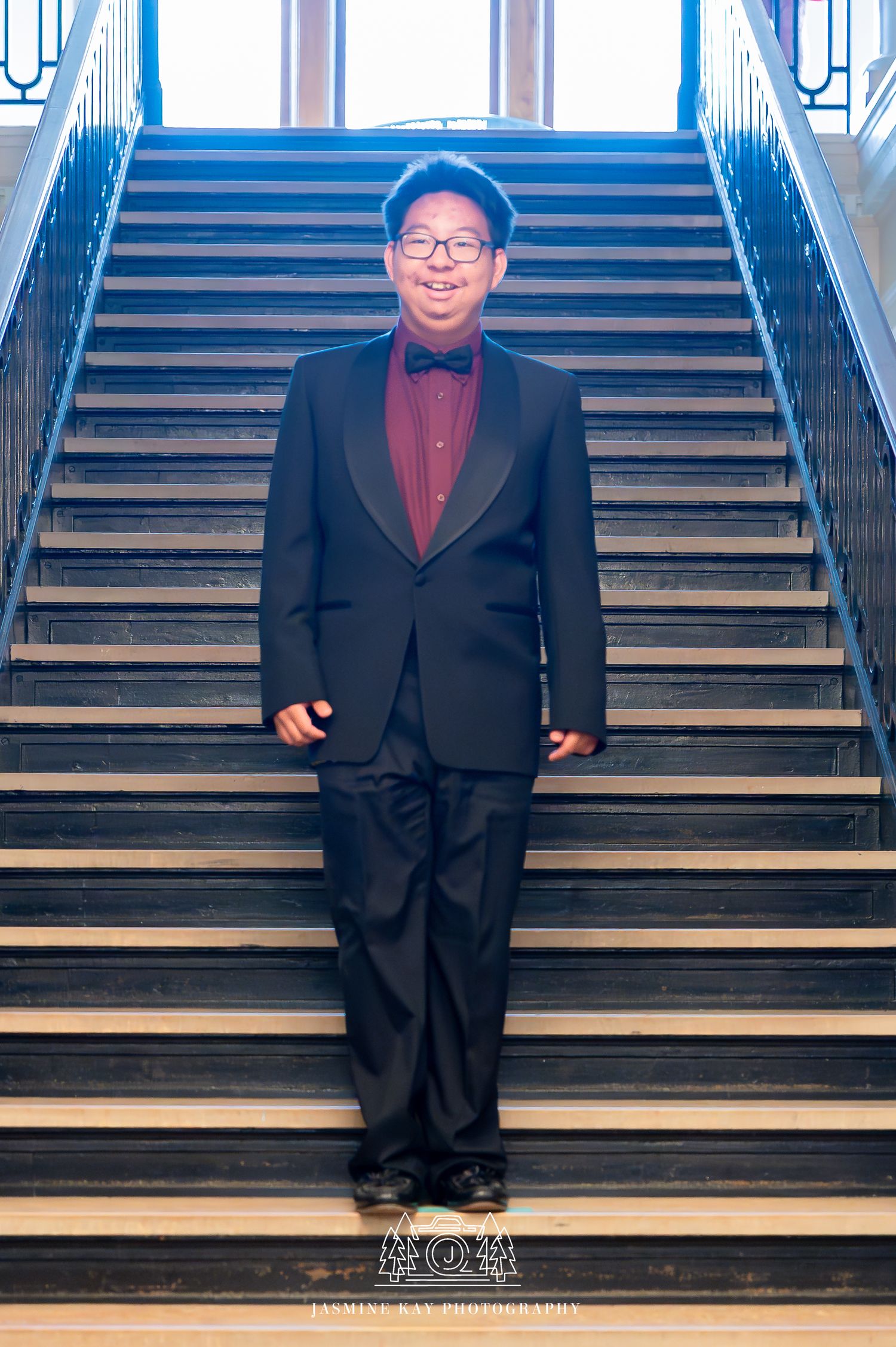 A person in a formal black tuxedo and bow tie stands on wooden stairs in front of bright blue-lit windows.