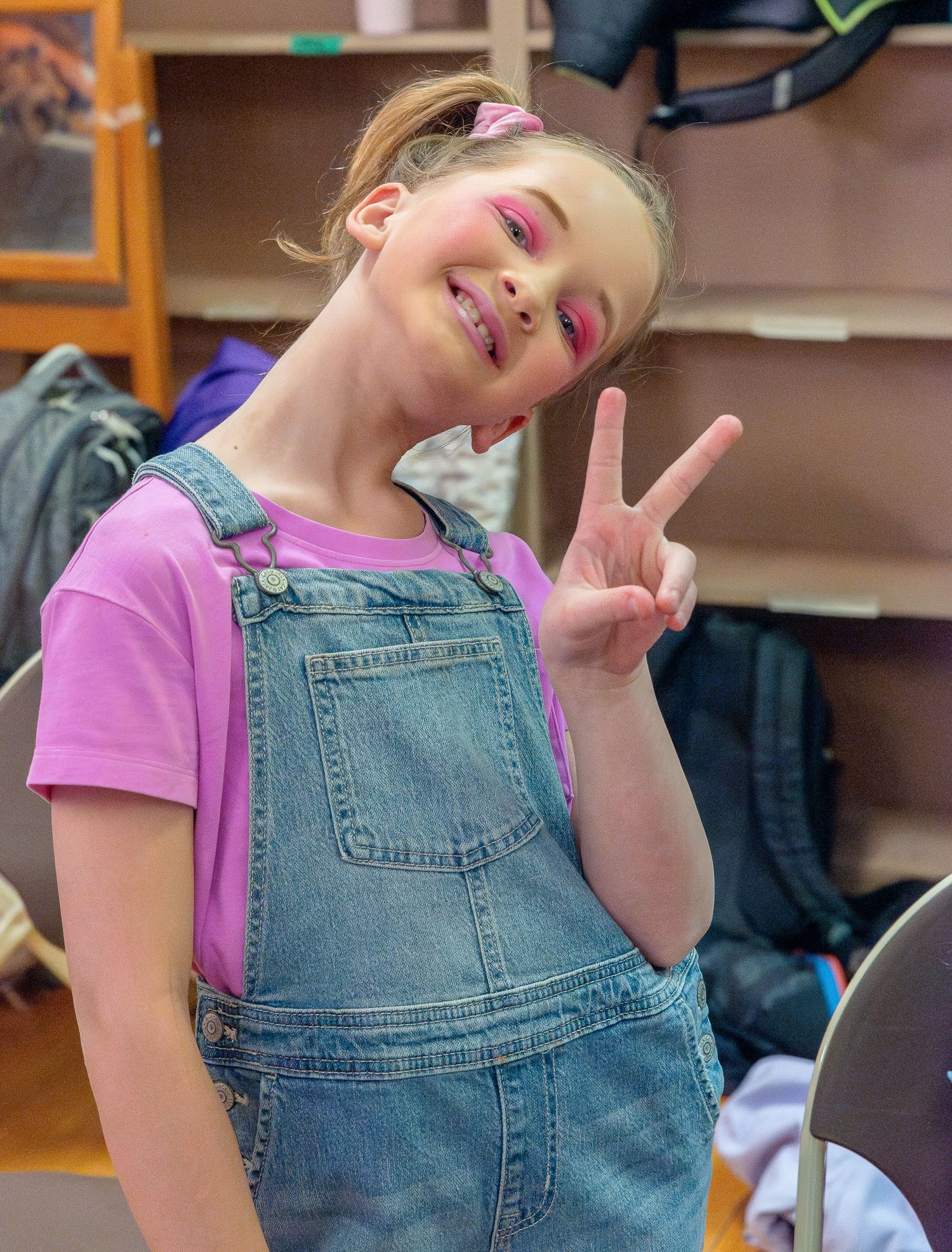 A performer in denim overalls and pink shirt making peace signs during a show.