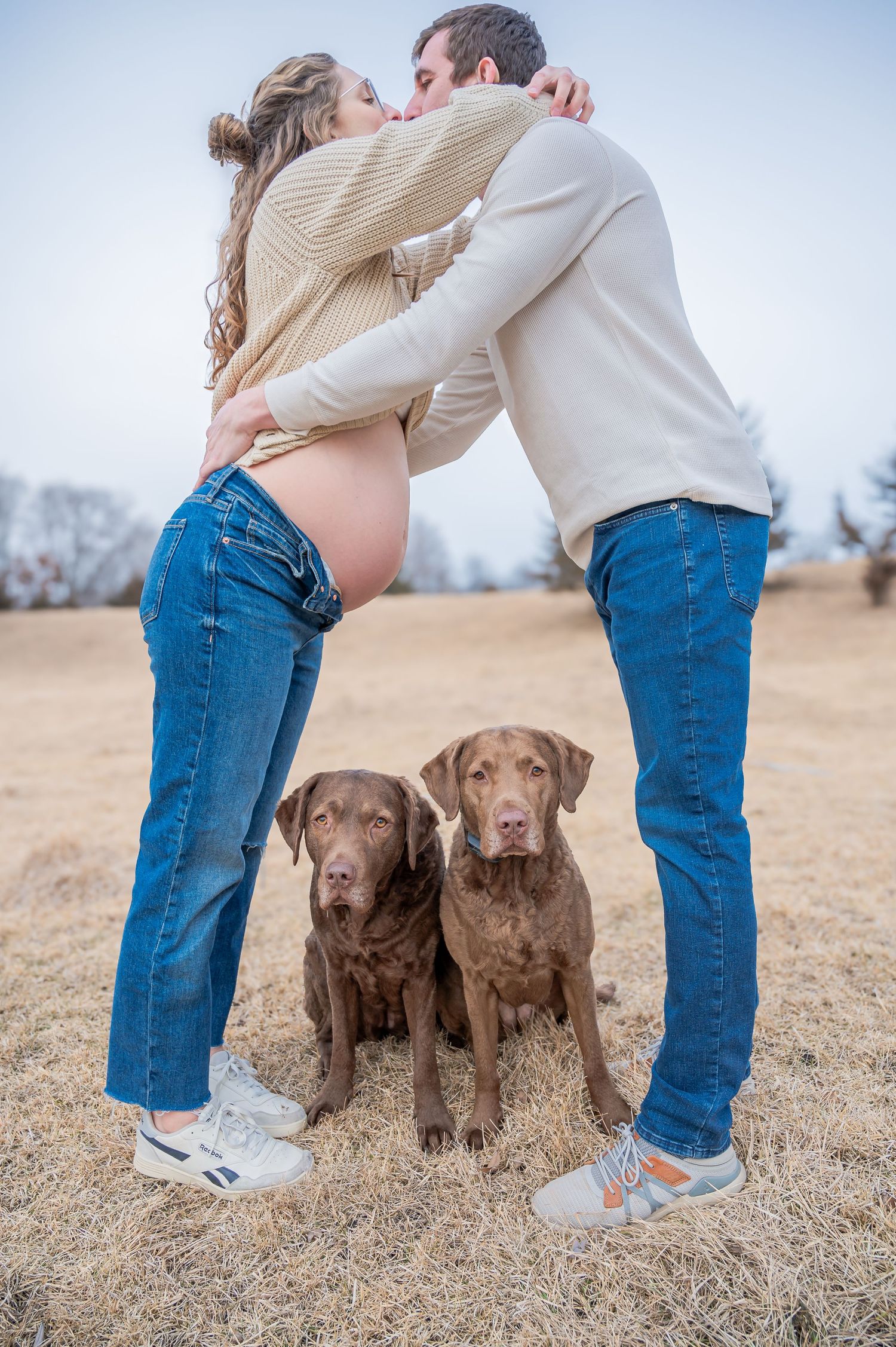 A couple embraces with their two Chesapeake Bay Retriever dogs sitting at their feet on dry grass.