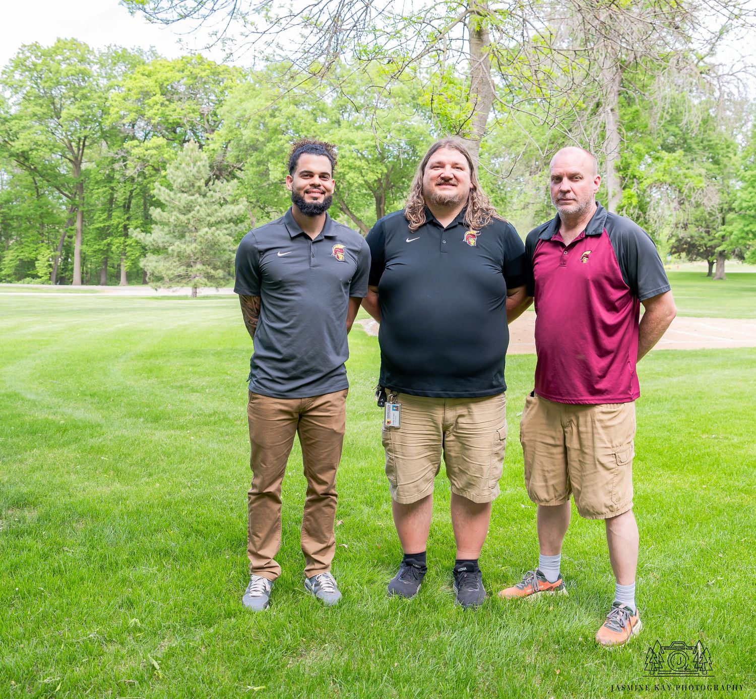 Three people stand together on a well-maintained green lawn with trees in the background on a sunny day.