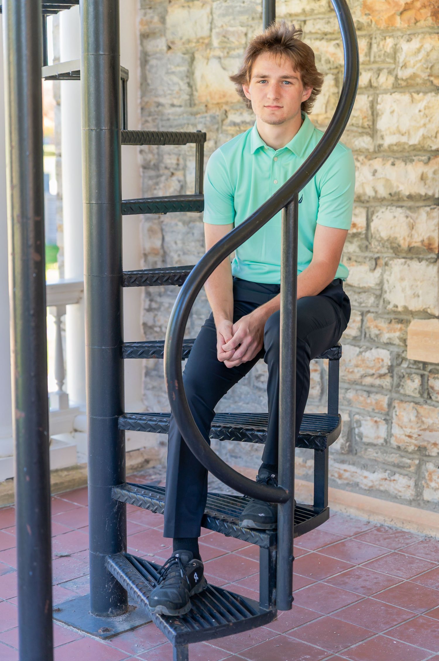 Person in turquoise shirt sits on steps against stone wall with black metal railings in architectural setting.