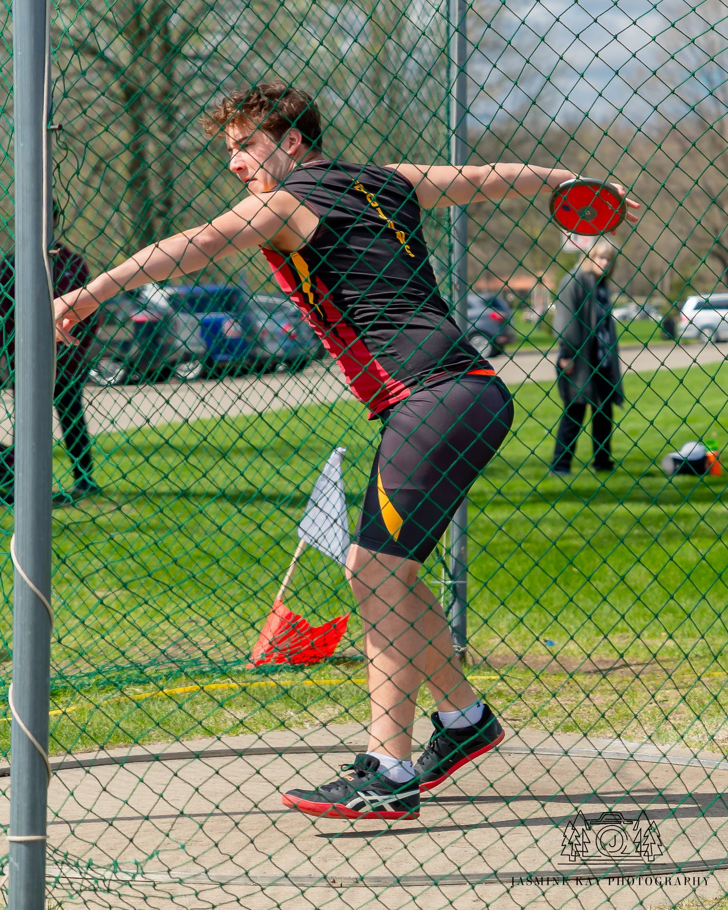 Athletes practicing discus throwing technique in an outdoor field with chain link fence in background.