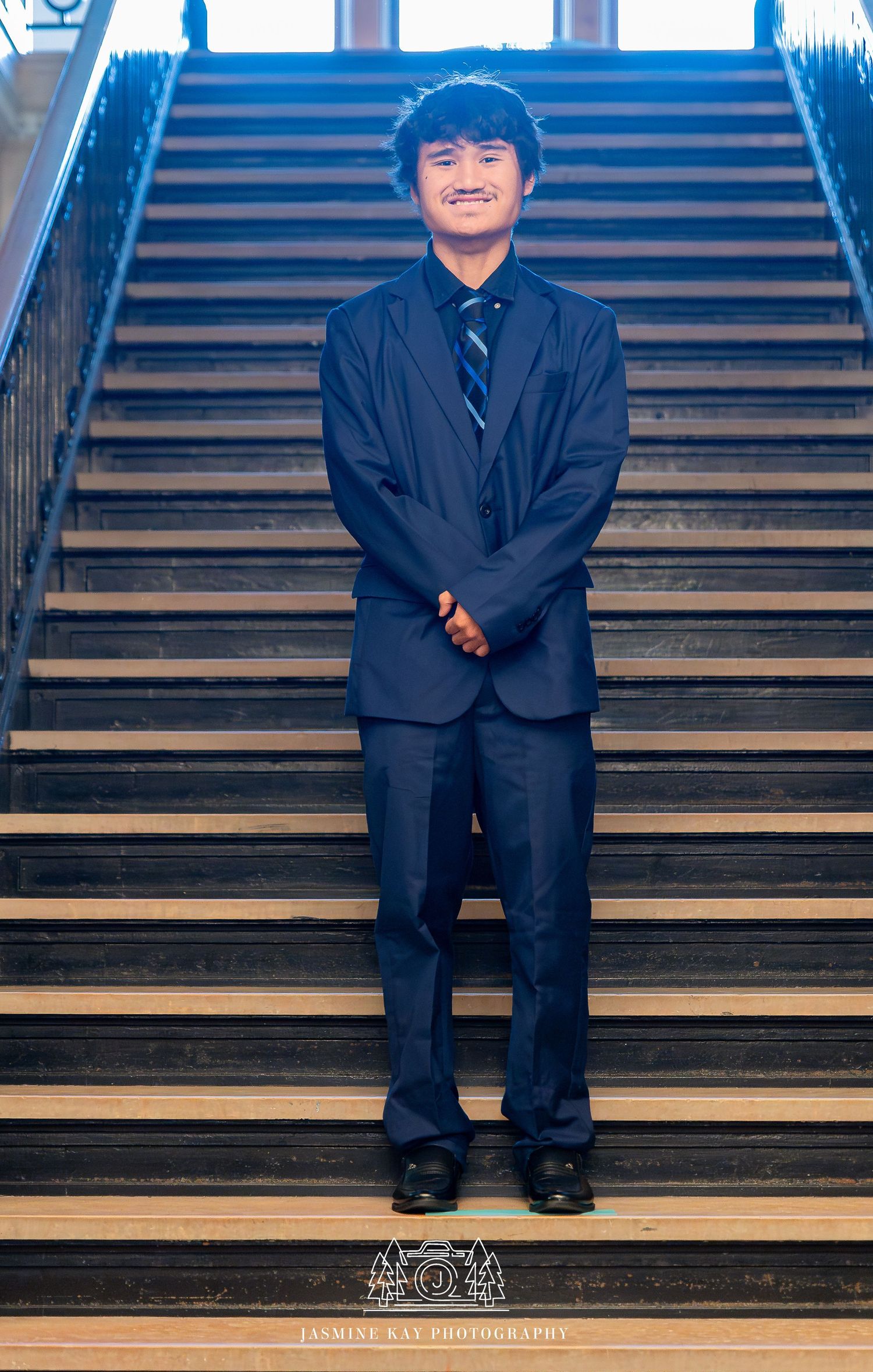 A person in a dark suit sits on wooden stairs with a bright blue background, creating a playful and casual pose.