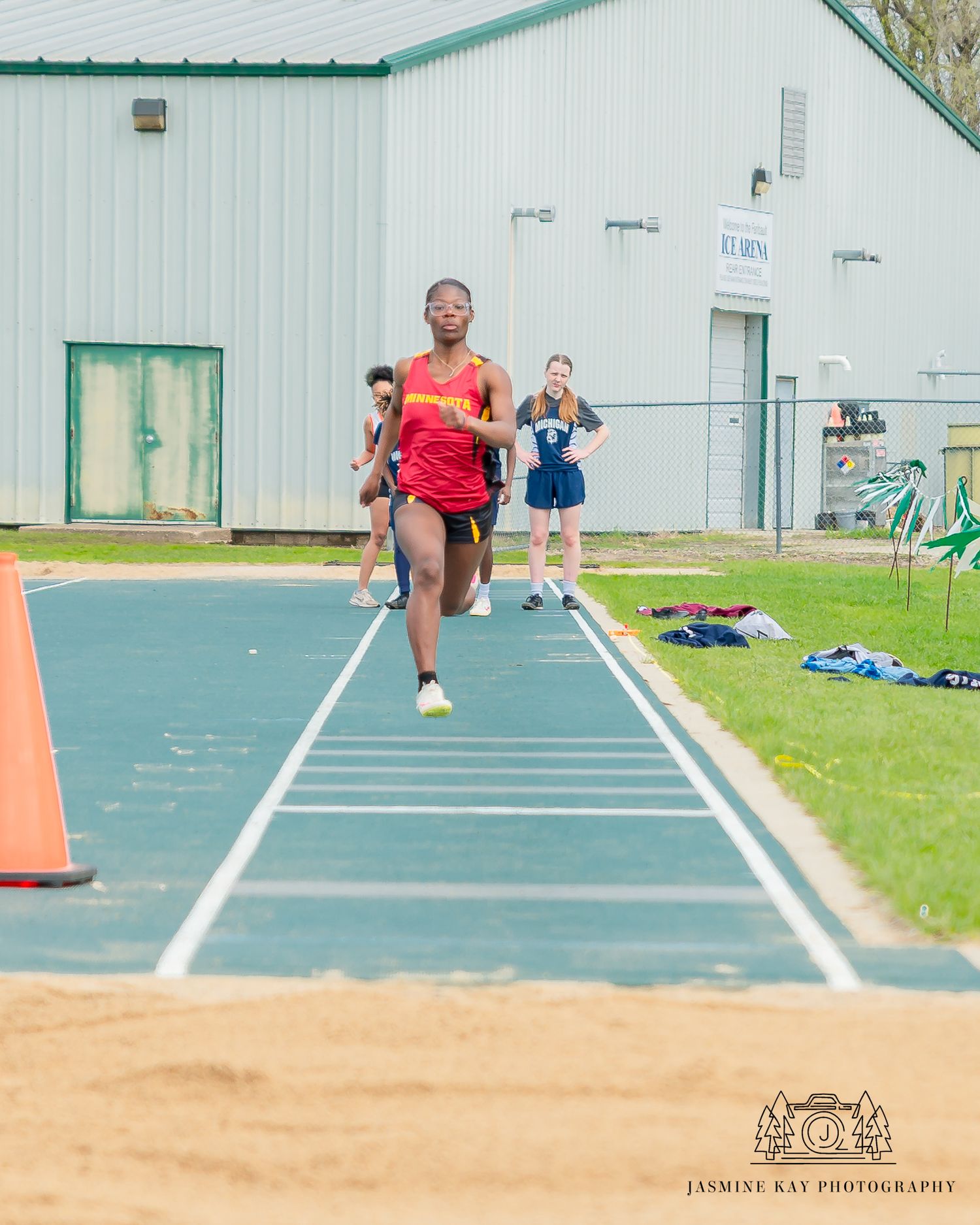 Long jumper in red uniform soars through the air over a sand pit during a track and field competition.