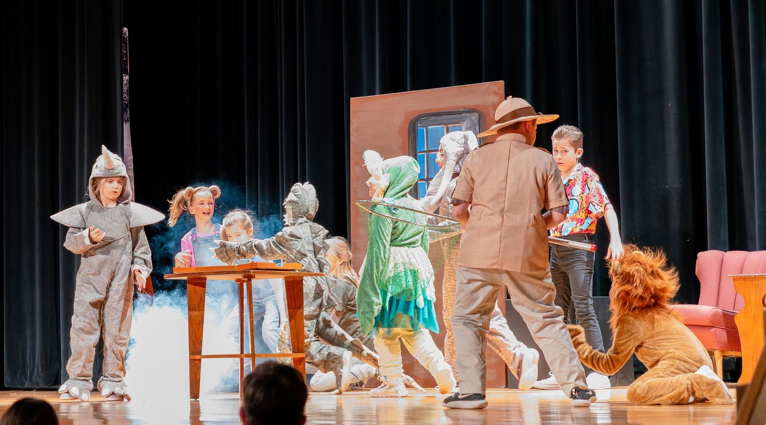 Theater actors perform a scene on stage with minimal props including wooden furniture.