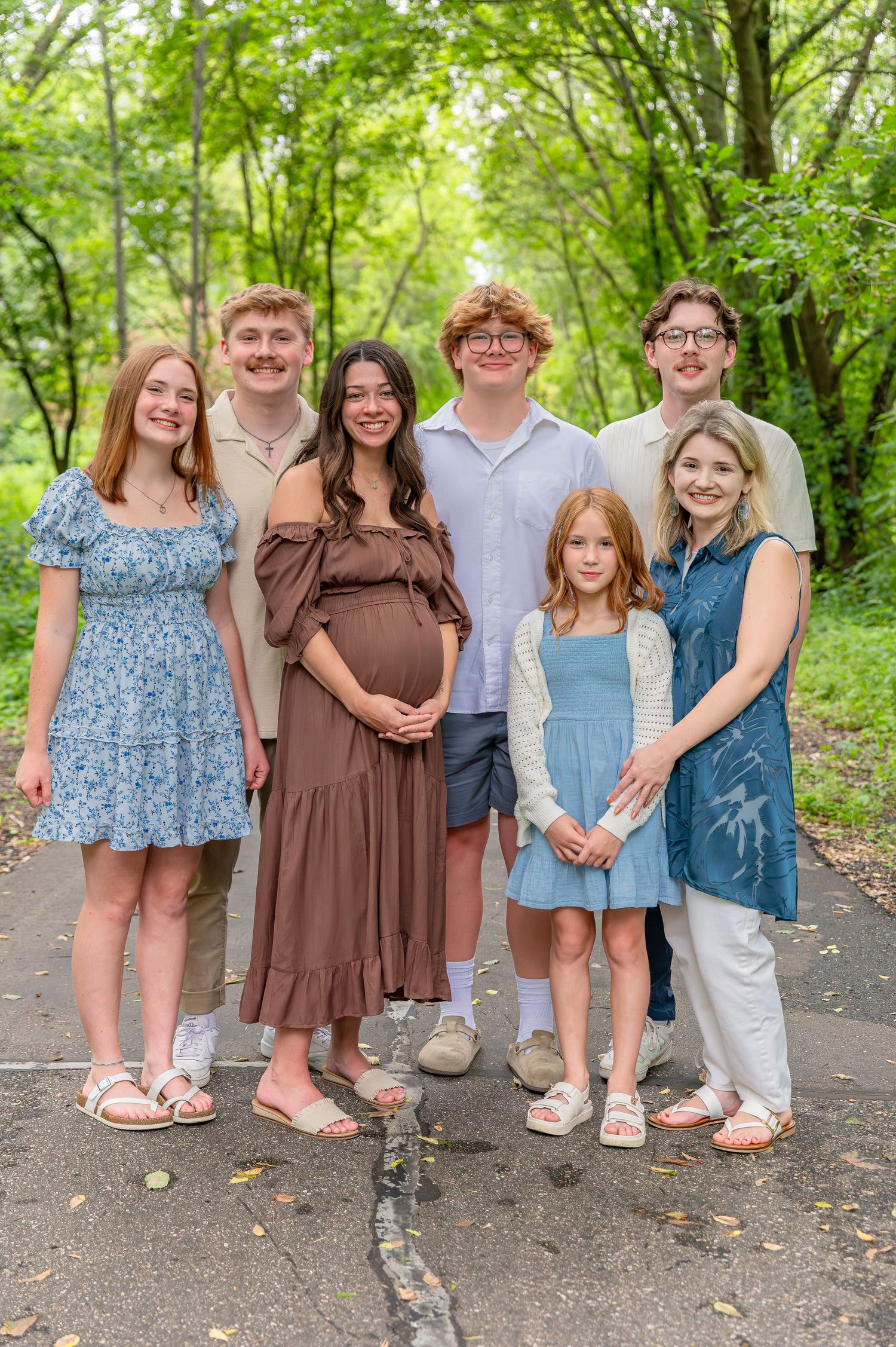 A group of children stand together on a path in a lush green forest setting wearing coordinated blue and brown outfits.