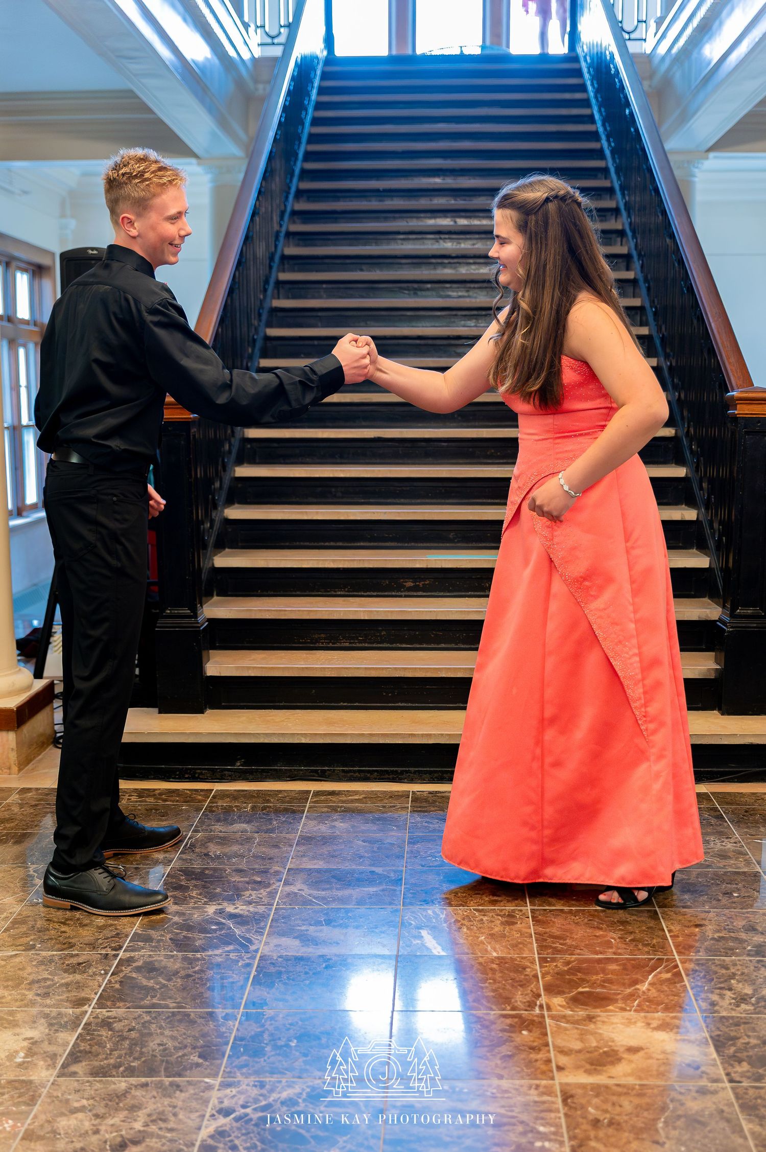 A couple in formal attire performs a romantic proposal on a grand marble staircase in an elegant indoor setting.