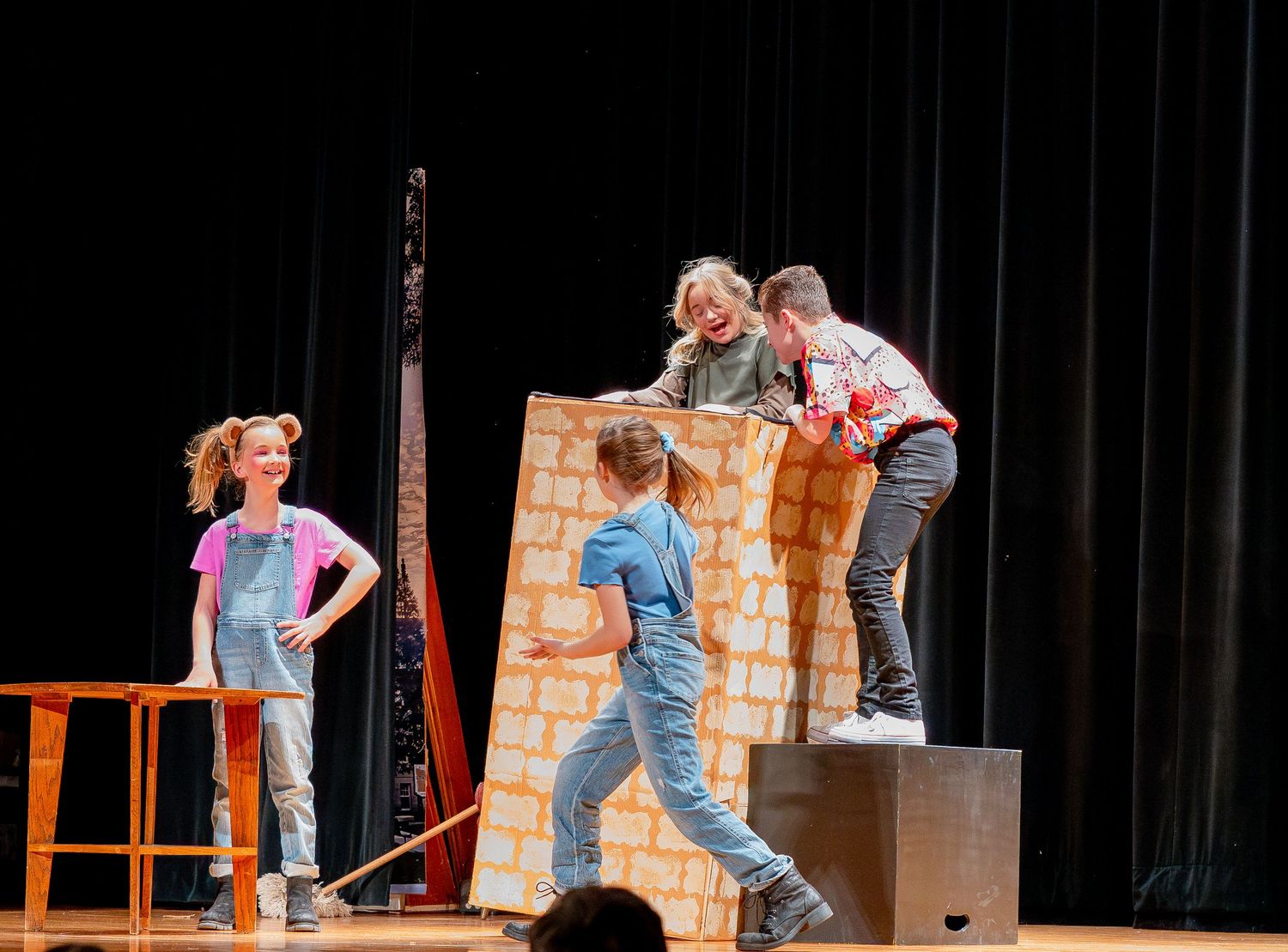 Theater performers acting on stage with cardboard props and scenery during a play performance.