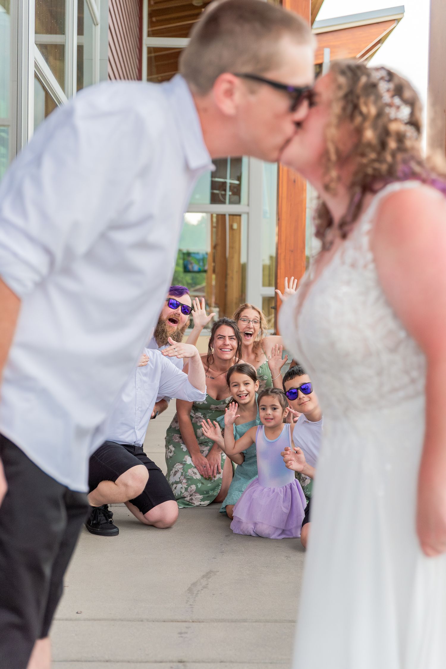 Playful wedding photo with onlookers reacting in background at wooden venue entrance.