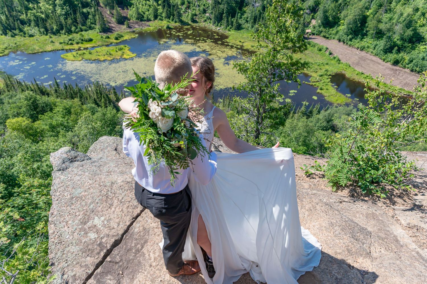 A romantic wedding photo taken on a scenic cliff overlook with a pond and forest landscape in the background.