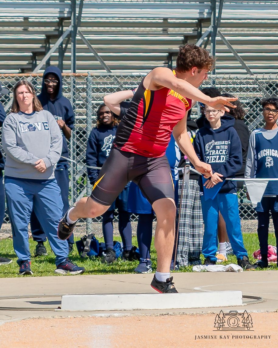Athletes compete in long jump event at outdoor track meet with spectators watching from bleachers in background.