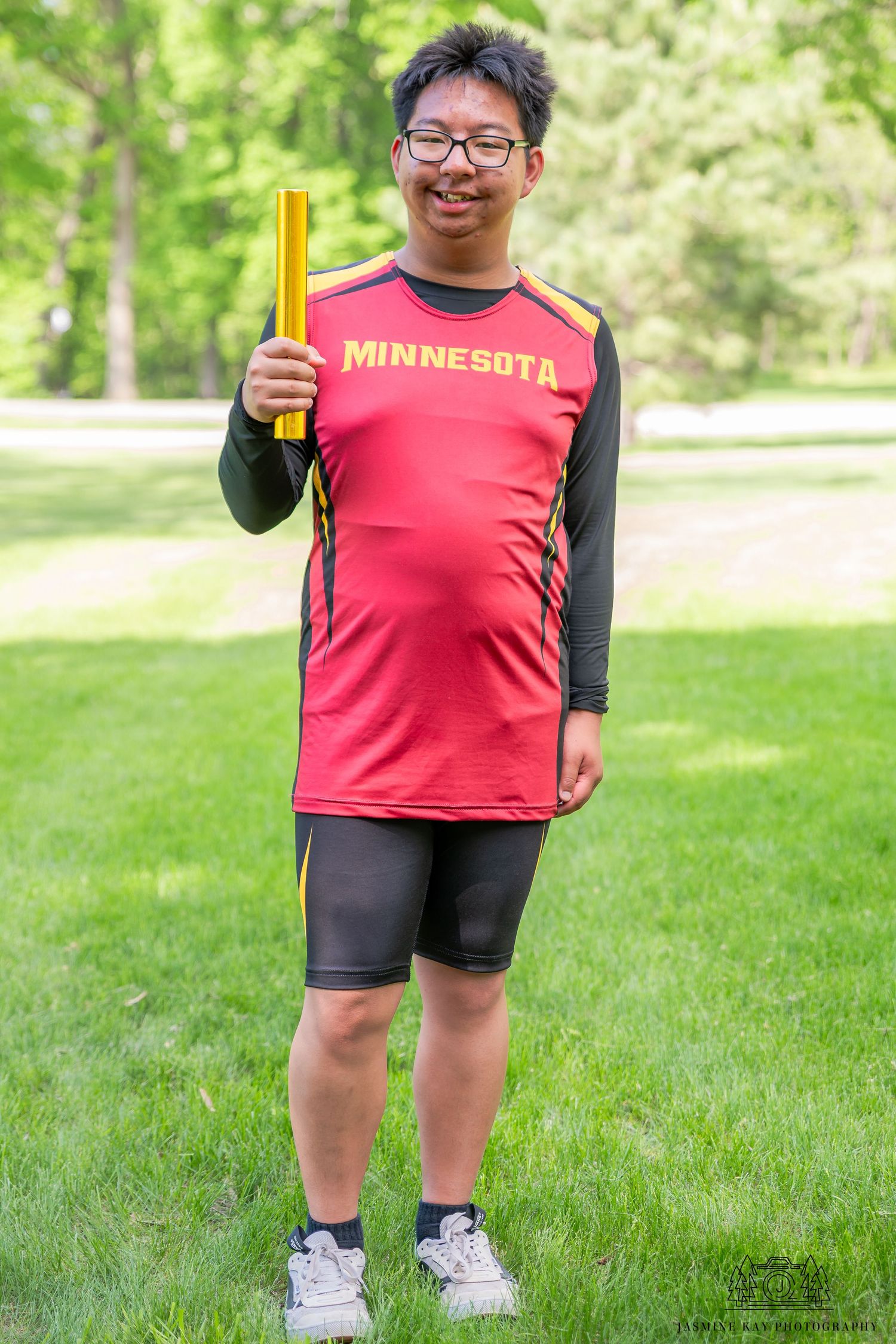 Person wearing red Minnesota shirt and black shorts holding a yellow baton stands on grass in a park setting.