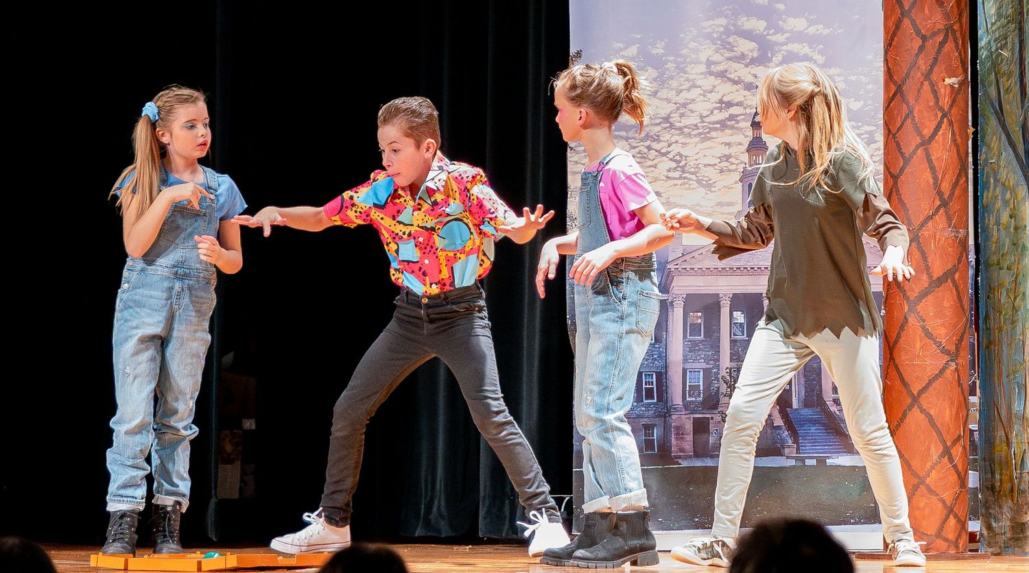 Theater actors engage in dramatic movement sequence on stage with rustic scenery and props.