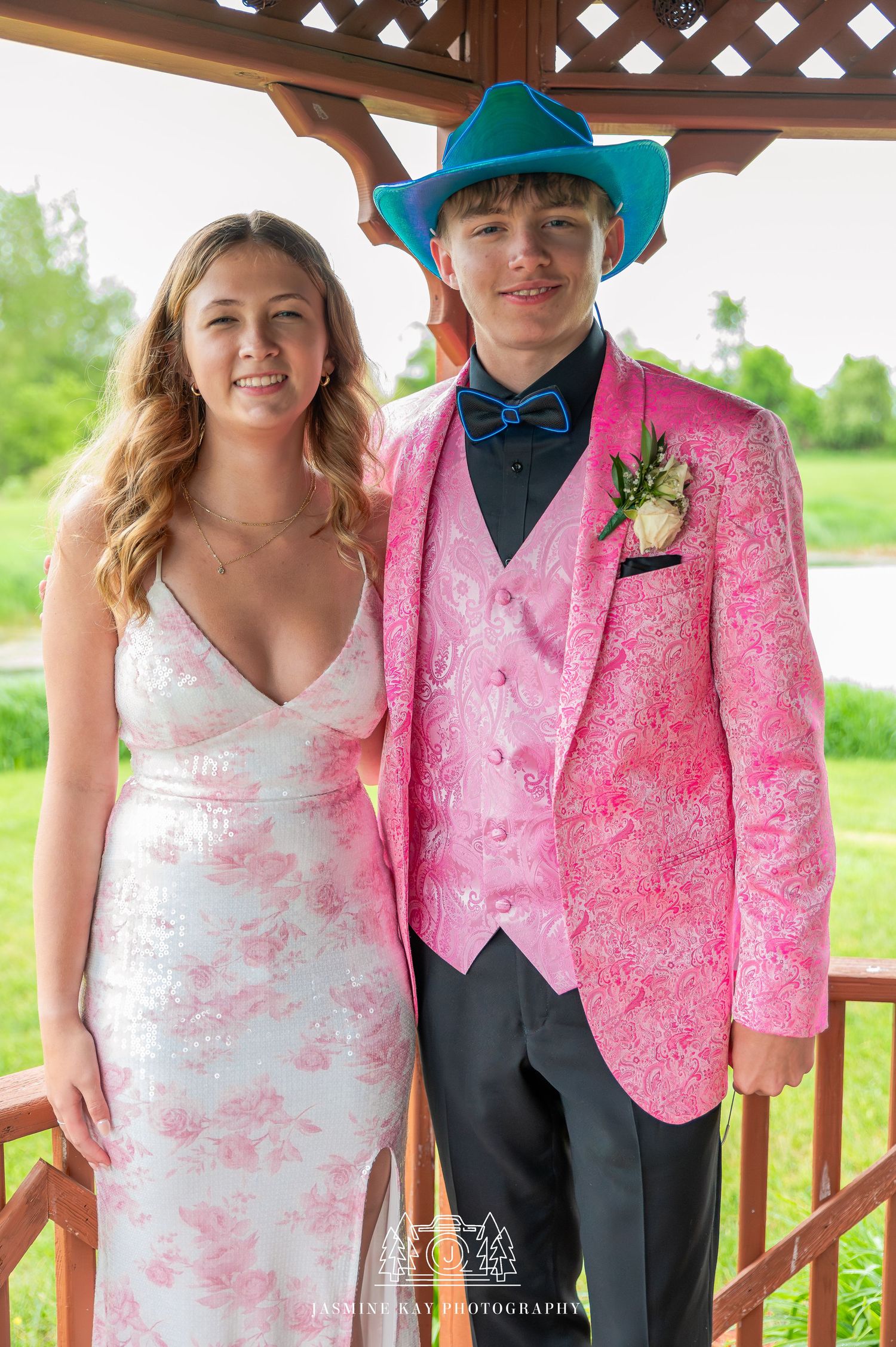 Couple in formal attire with pink and turquoise accents pose together on a wooden deck with greenery in background.
