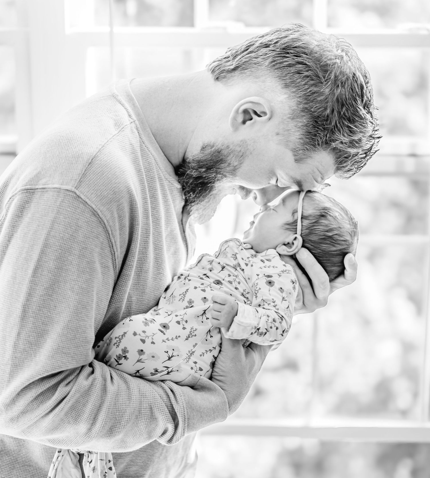 A tender black and white portrait of a bearded parent gently kissing a baby wearing a floral onesie.