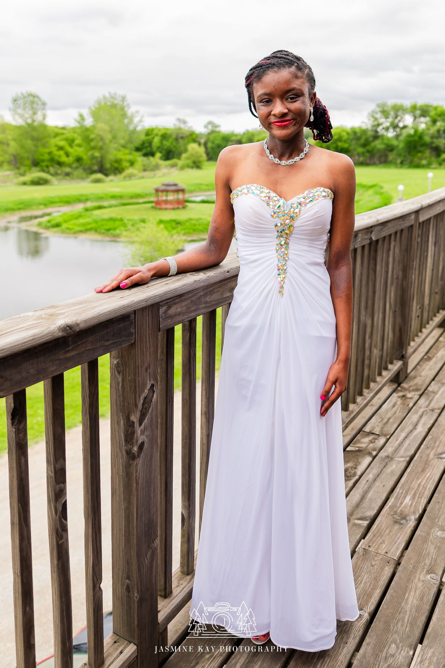 Elegant white strapless gown with gold embellishments photographed on a wooden deck overlooking a serene pond.