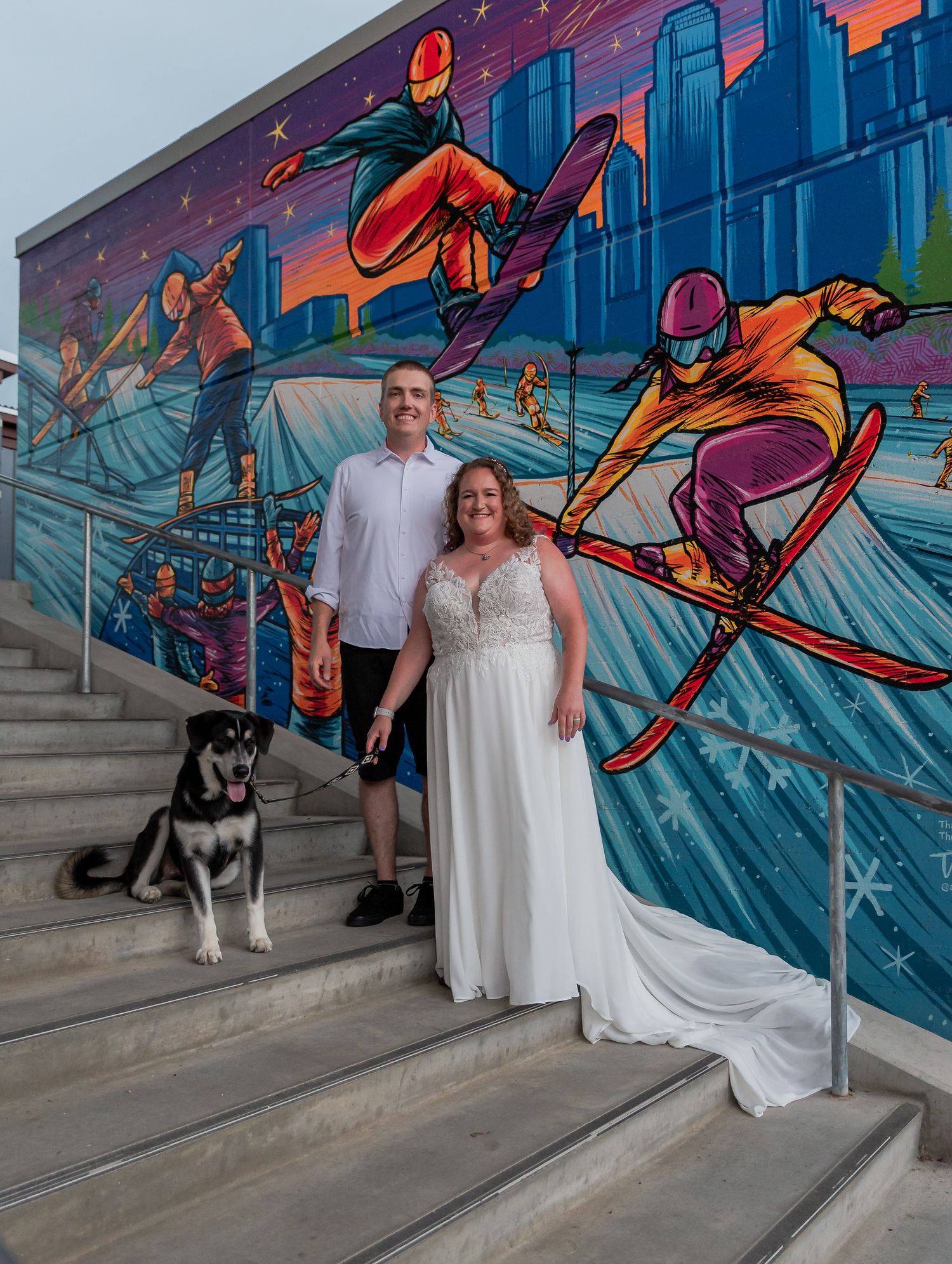 A couple poses with their black and white dog against a vibrant blue graffiti mural on concrete steps.