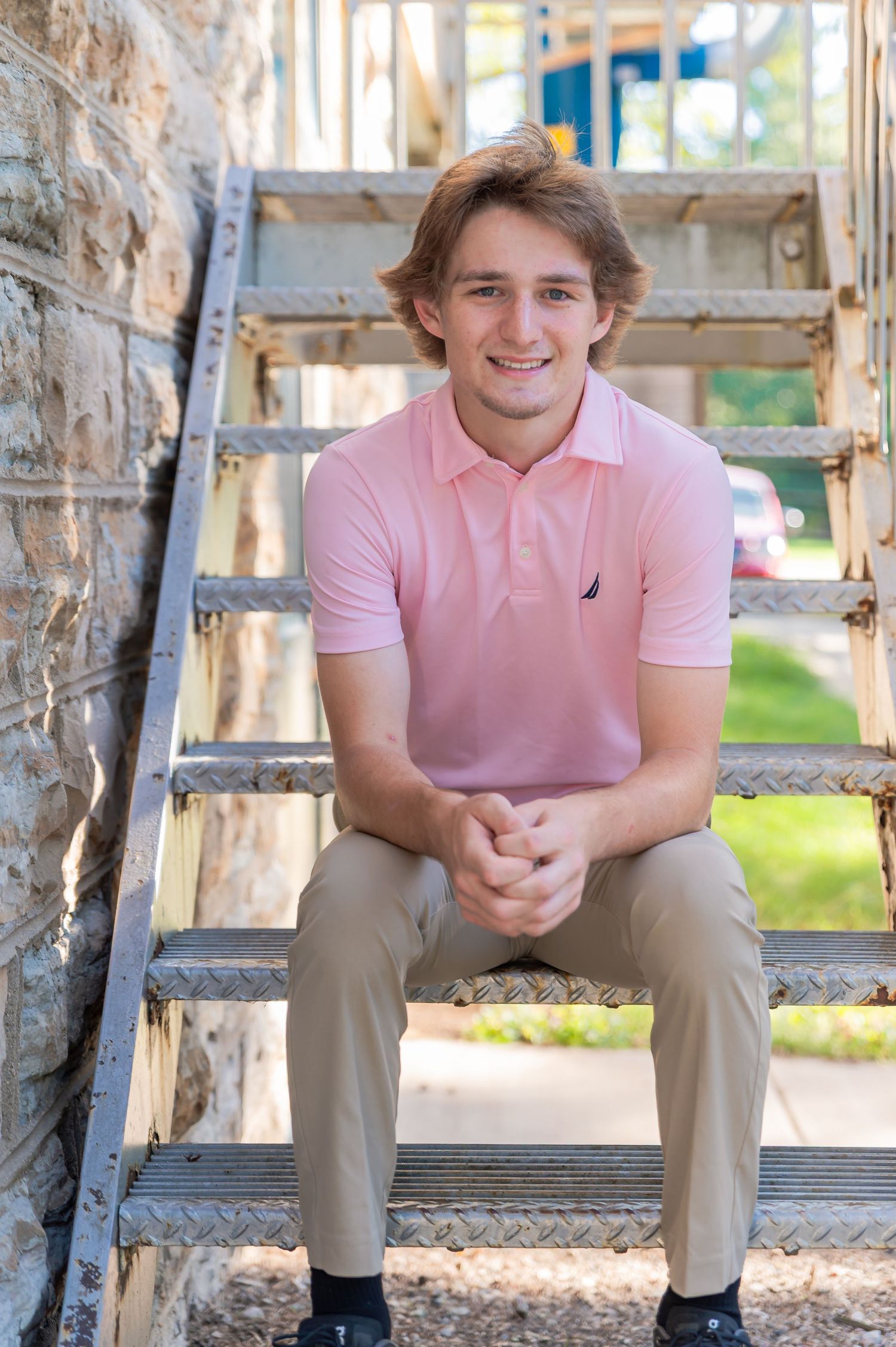 A person in a pink polo shirt and khaki pants sits casually on rustic wooden steps in an outdoor setting.