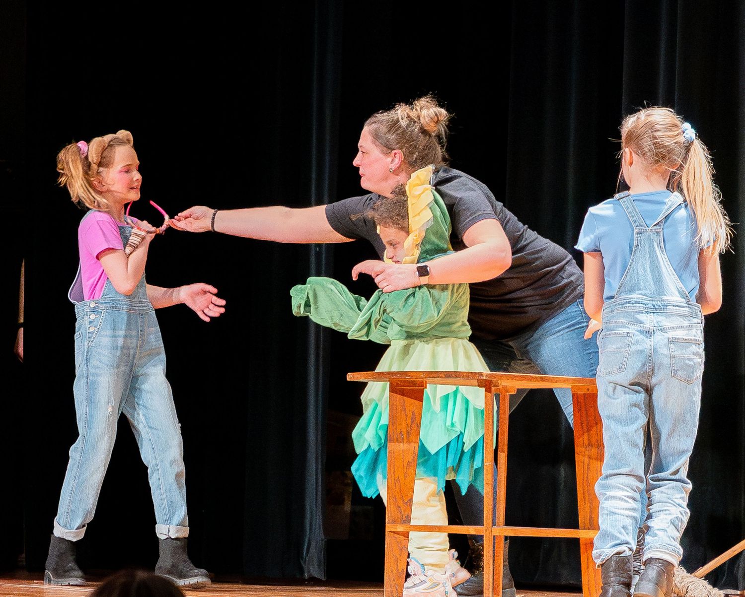 Performers act out a dynamic scene on stage with wooden table props against a brick wall set.