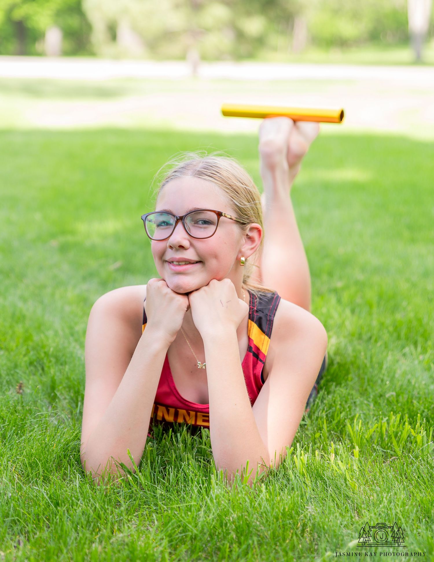 Student in glasses and athletic wear lying on grass holding a yellow relay baton during track practice.