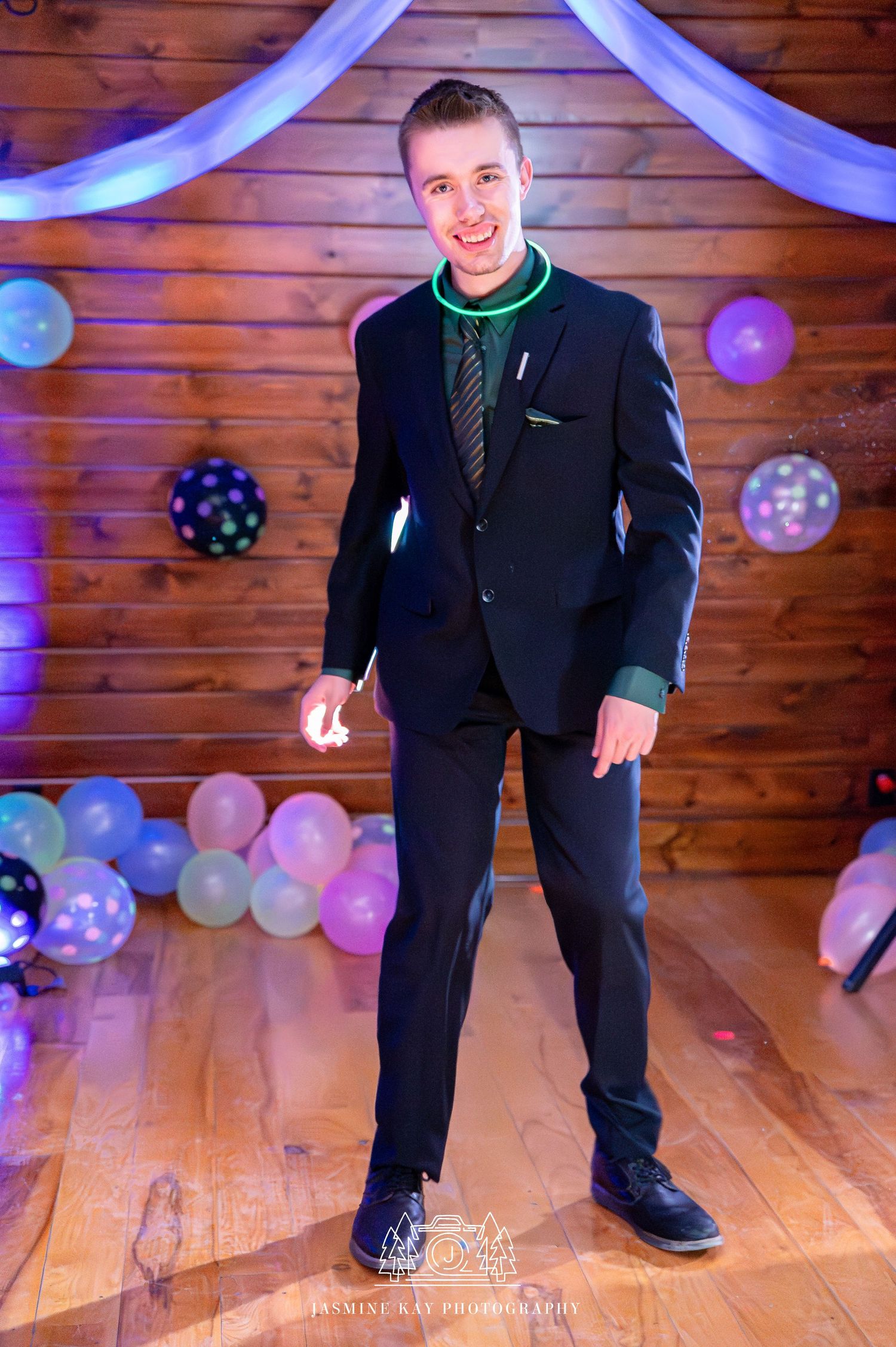 Person in formal black suit poses playfully on wooden dance floor with blue and pink balloon decorations.