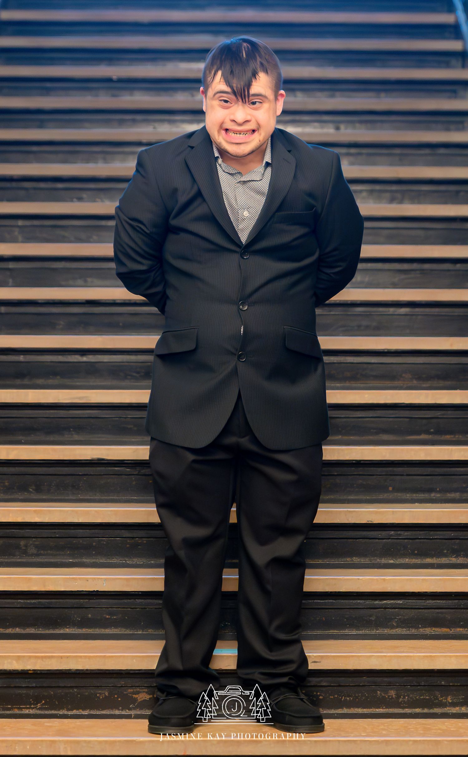 A person in a black suit and gray shirt stands smiling on wooden stairs.
