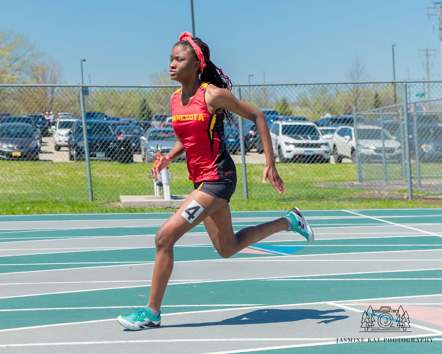 Athlete sprinting on outdoor track wearing red uniform and turquoise shoes.