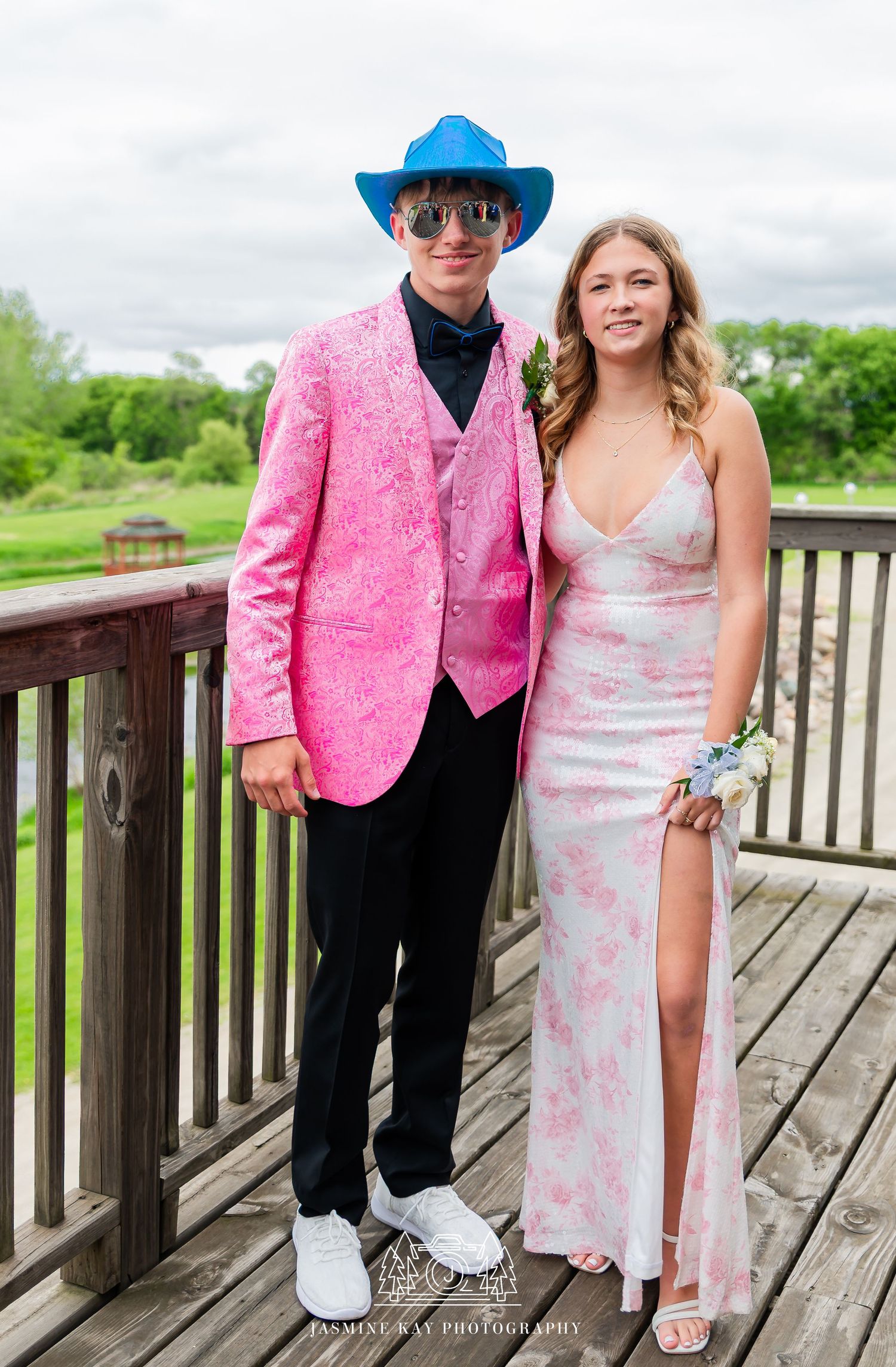 A couple poses for prom photos on a wooden deck, wearing a bright pink jacket and white floral dress.