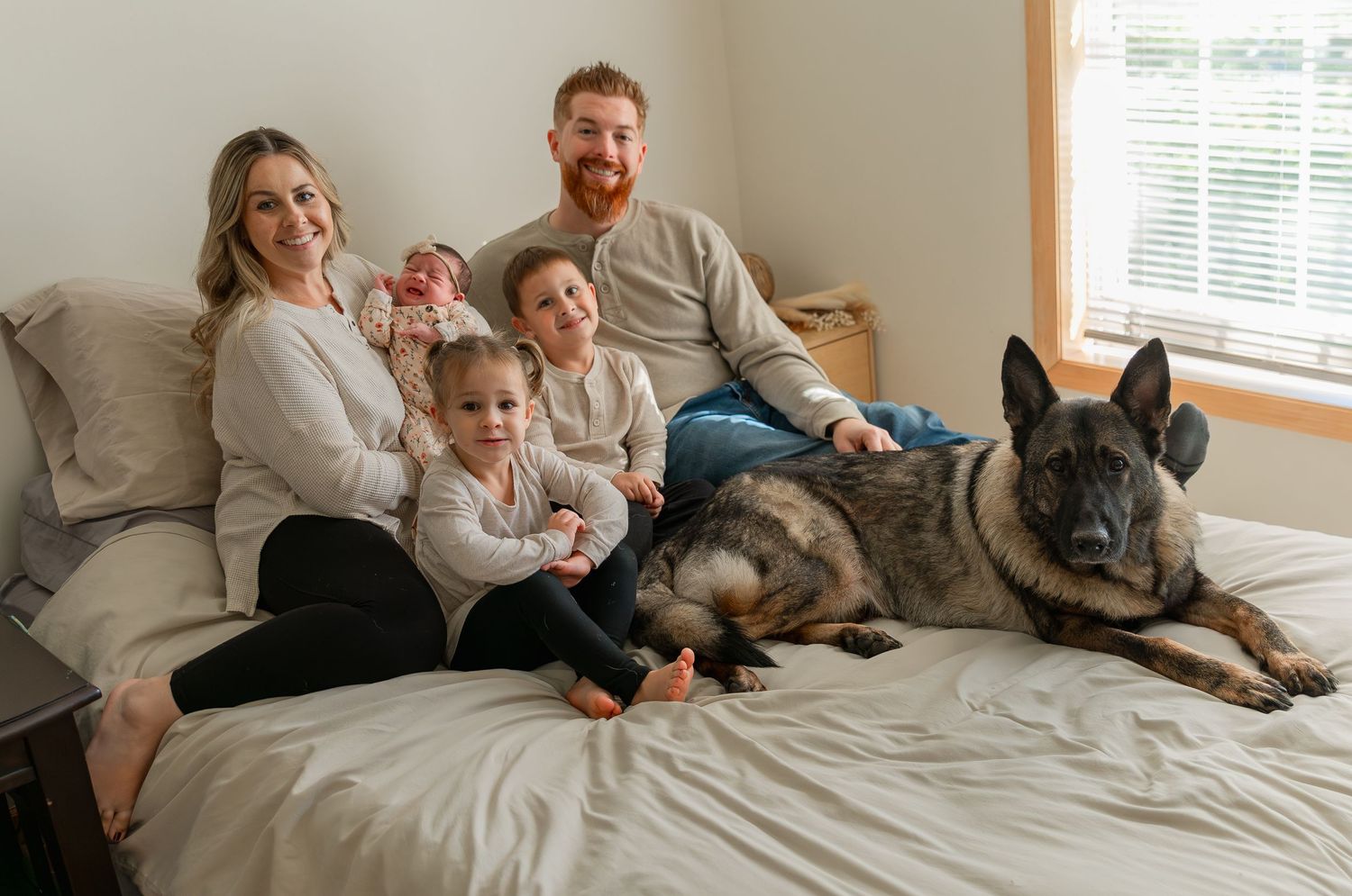 Family relaxing together on white bed with their dog in sunlit bedroom.