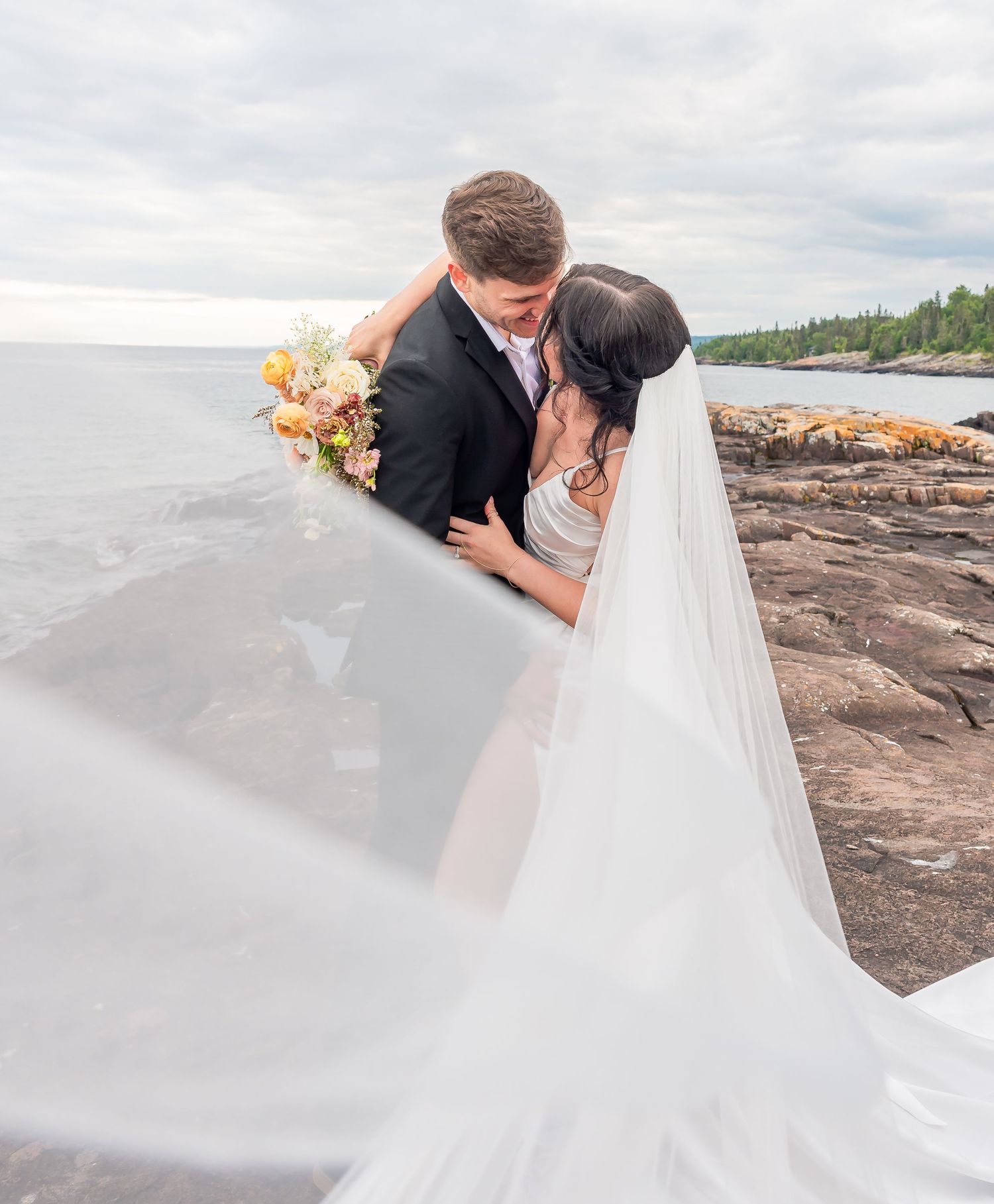 Wedding couple shares intimate moment on rocky shore with flowing veil and peach bouquet.