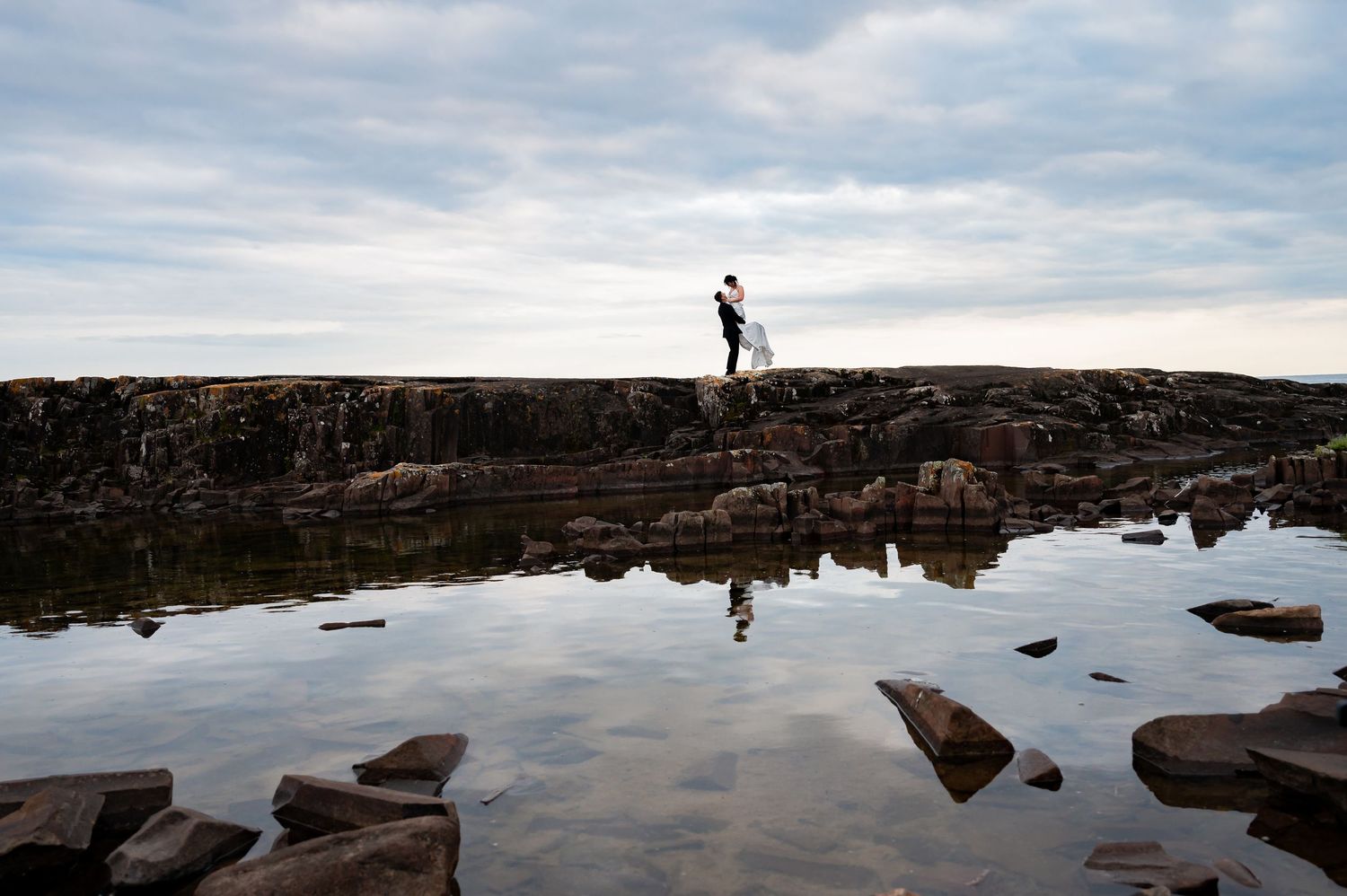 A silhouetted figure stands on rocky cliffs above still water reflecting clouds and stone formations at sunset.