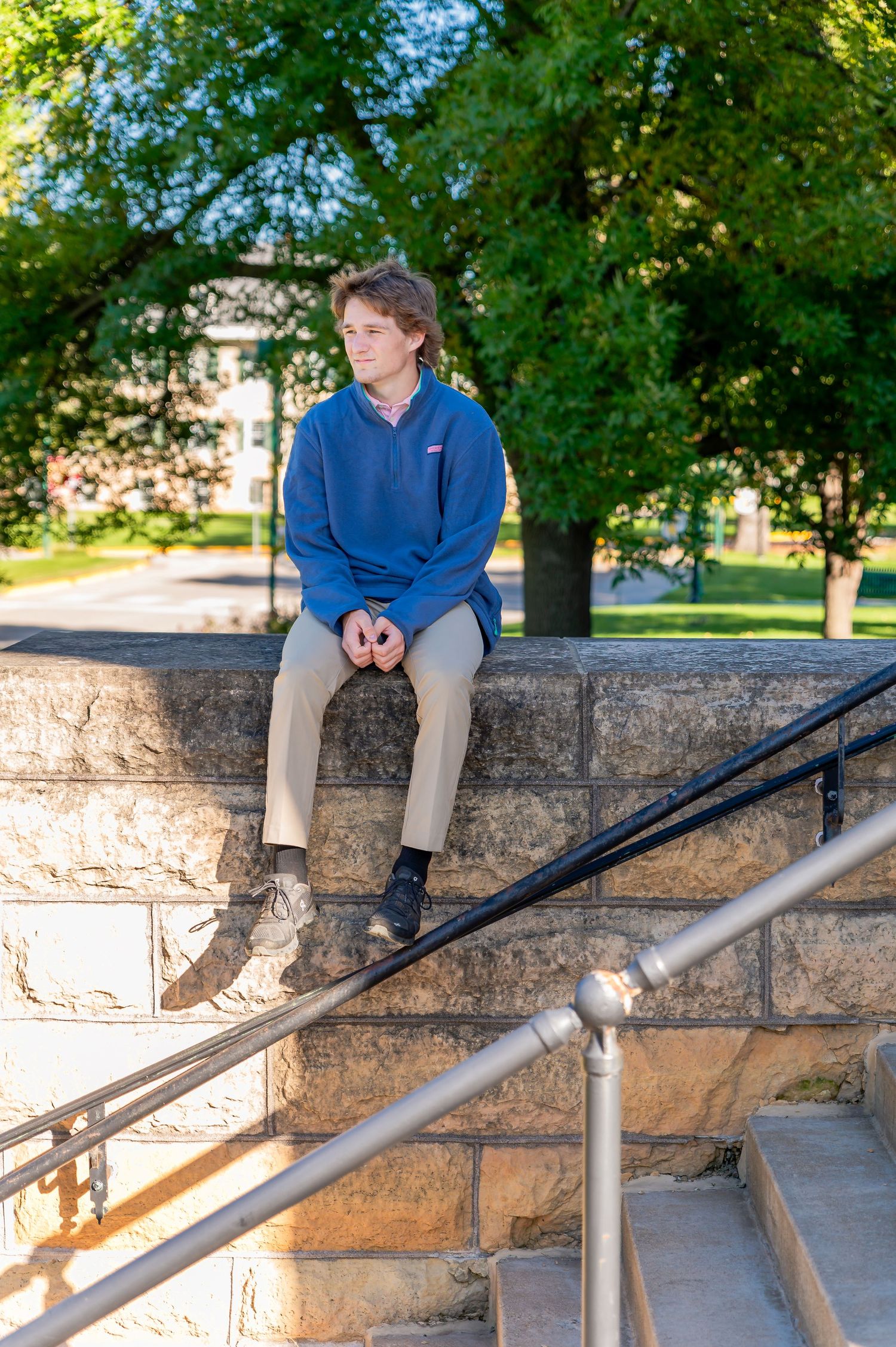 Someone in a blue sweater sits on a stone wall in a park setting with green trees in the background.