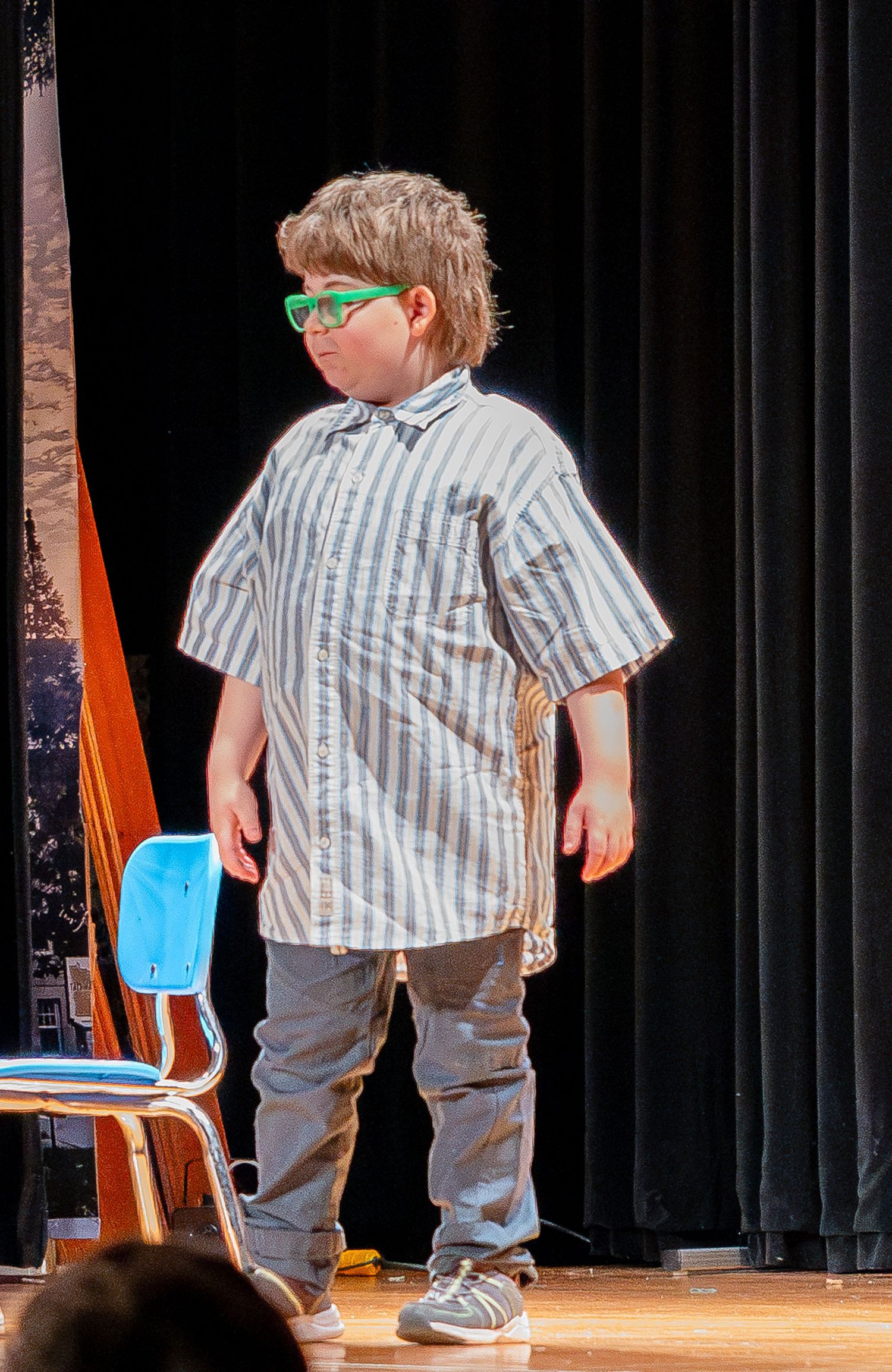 Student performer wearing bright green glasses, striped shirt and jeans stands on stage during a school production.