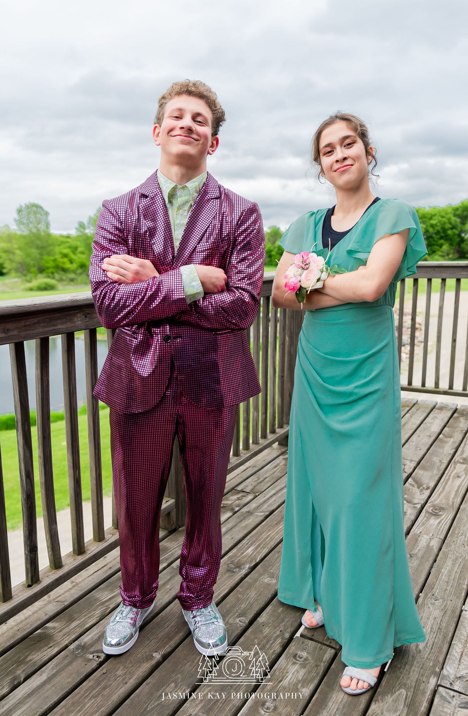 Two people in contrasting purple and teal outfits pose playfully on a wooden deck with green scenery in background.