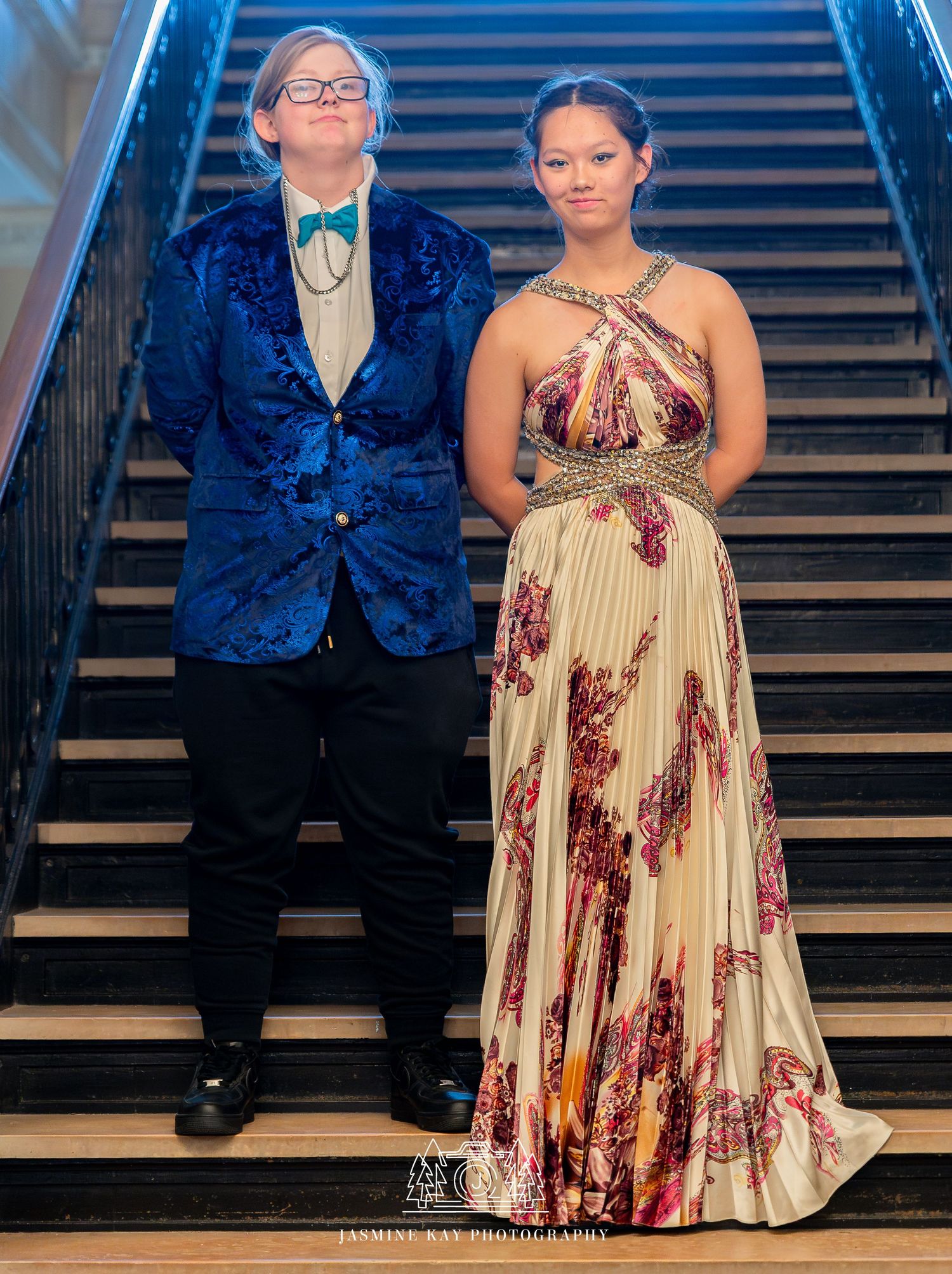 A couple in formal attire poses on a grand staircase, with the person wearing a floral print dress and navy suit.