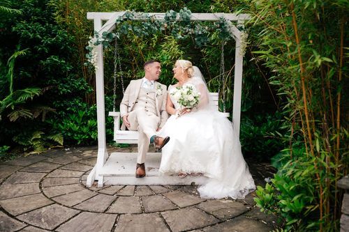 A couple sits together under a white garden arbor surrounded by lush greenery and circular stone patio.