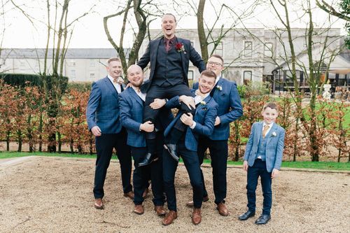 Groomsmen in navy suits playfully lift and pose together in a garden setting on a cloudy day.