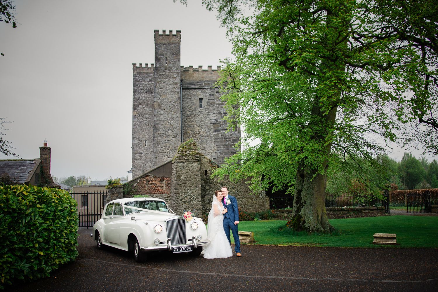 Classic white vintage car parked in front of a stone castle tower on a wedding day.