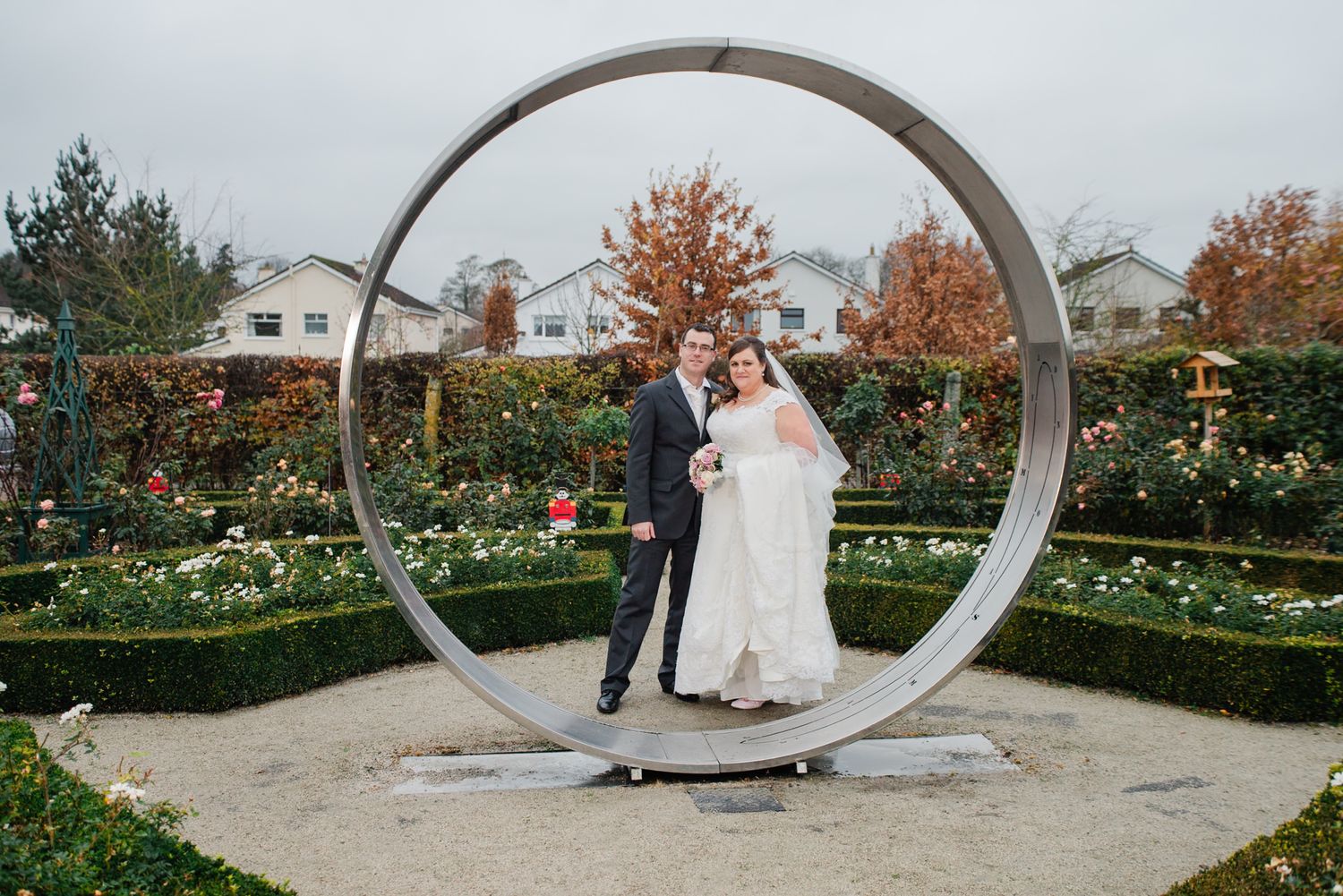 Wedding couple poses inside large circular metal sculpture in garden setting.