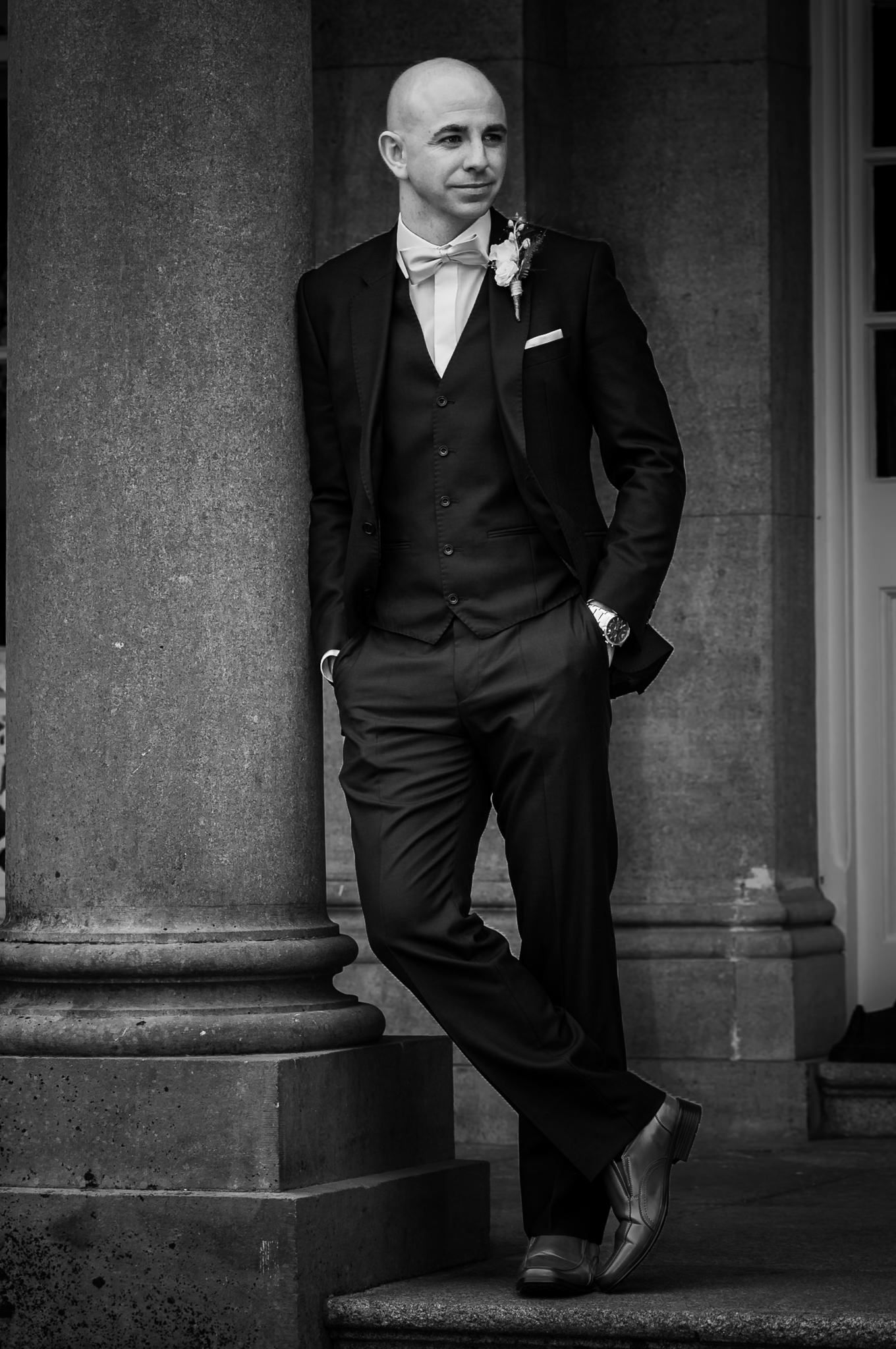 Black and white portrait of a groom in formal tuxedo with bowtie and boutonniere leaning against a stone column.