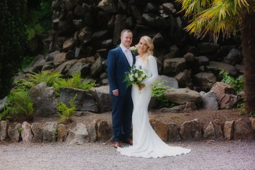 A couple stands together against a stone wall backdrop with tropical greenery in an outdoor wedding setting.