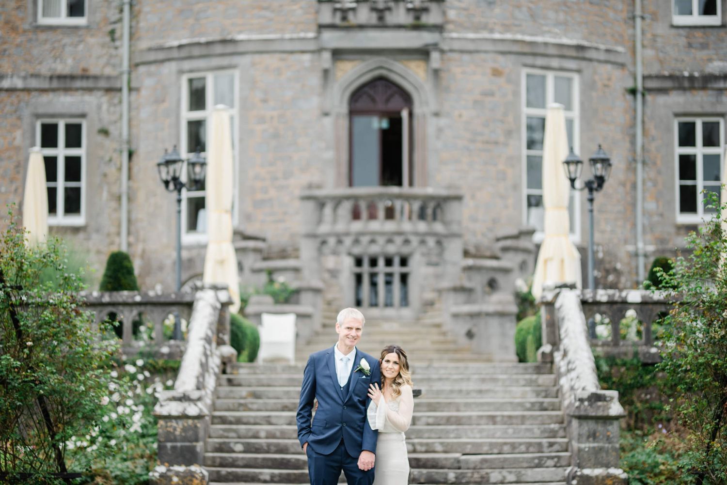 A couple stand on the grand stone staircase of a historic manor house flanked by statues and ornate architecture.