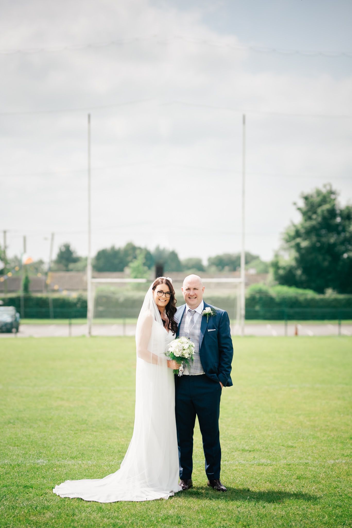 Wedding couple poses on sports field with goal posts in background.
